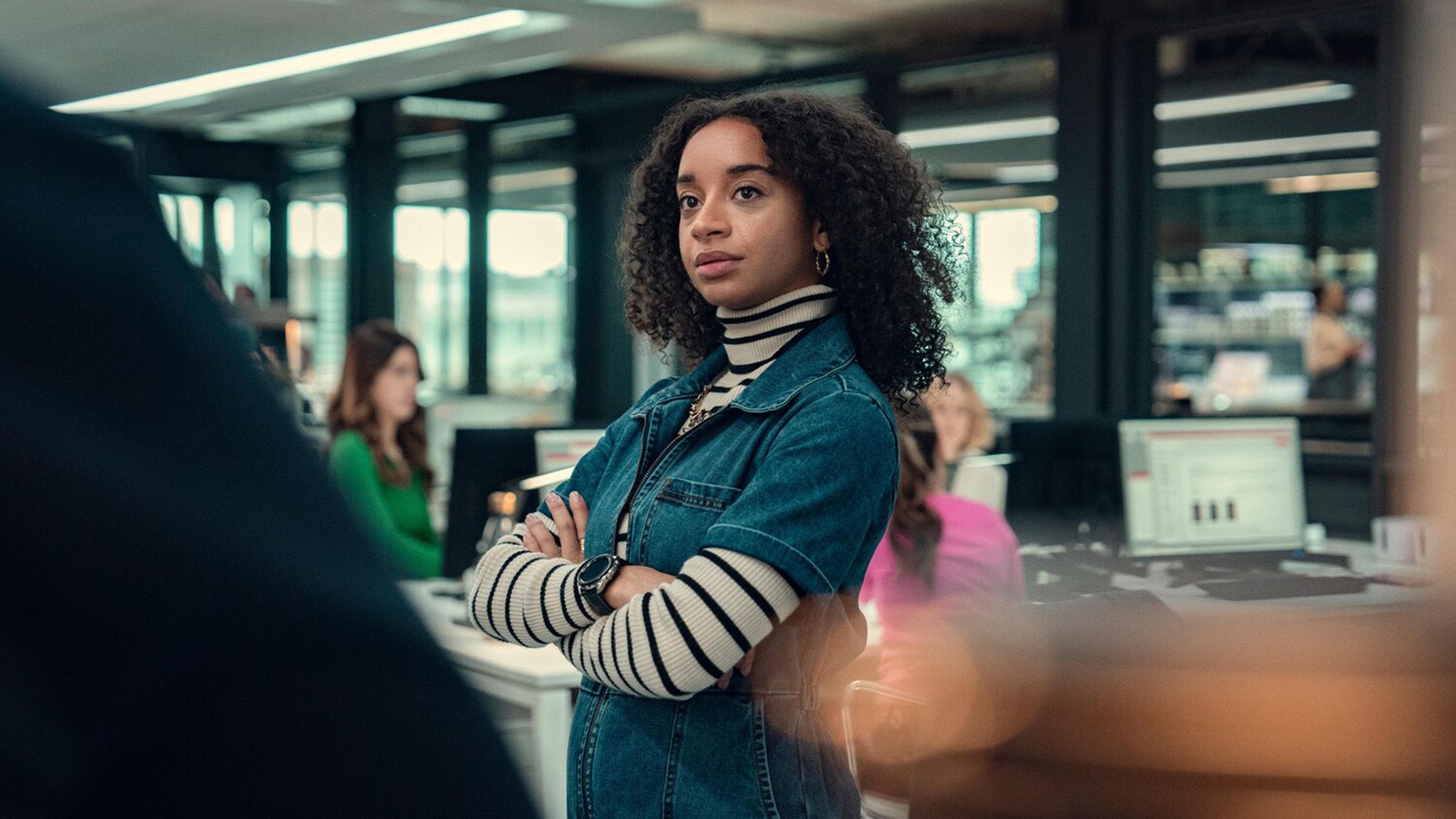 Siena Kelly con un maglione a righe e una camicia di jeans sta in mezzo all'ufficio, con le braccia incrociate sul petto