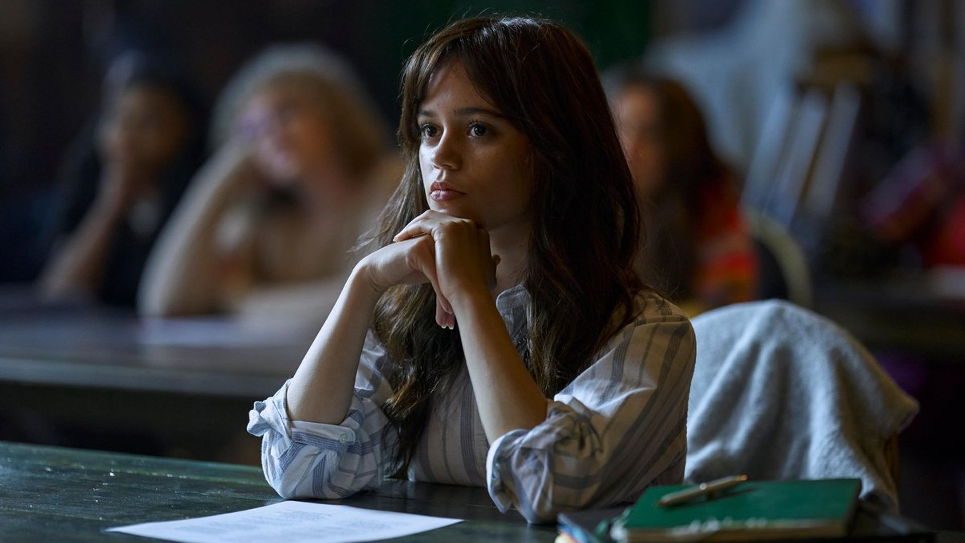 Jenna Ortega in un maglione bianco seduta al banco durante la lezione con un'espressione concentrata