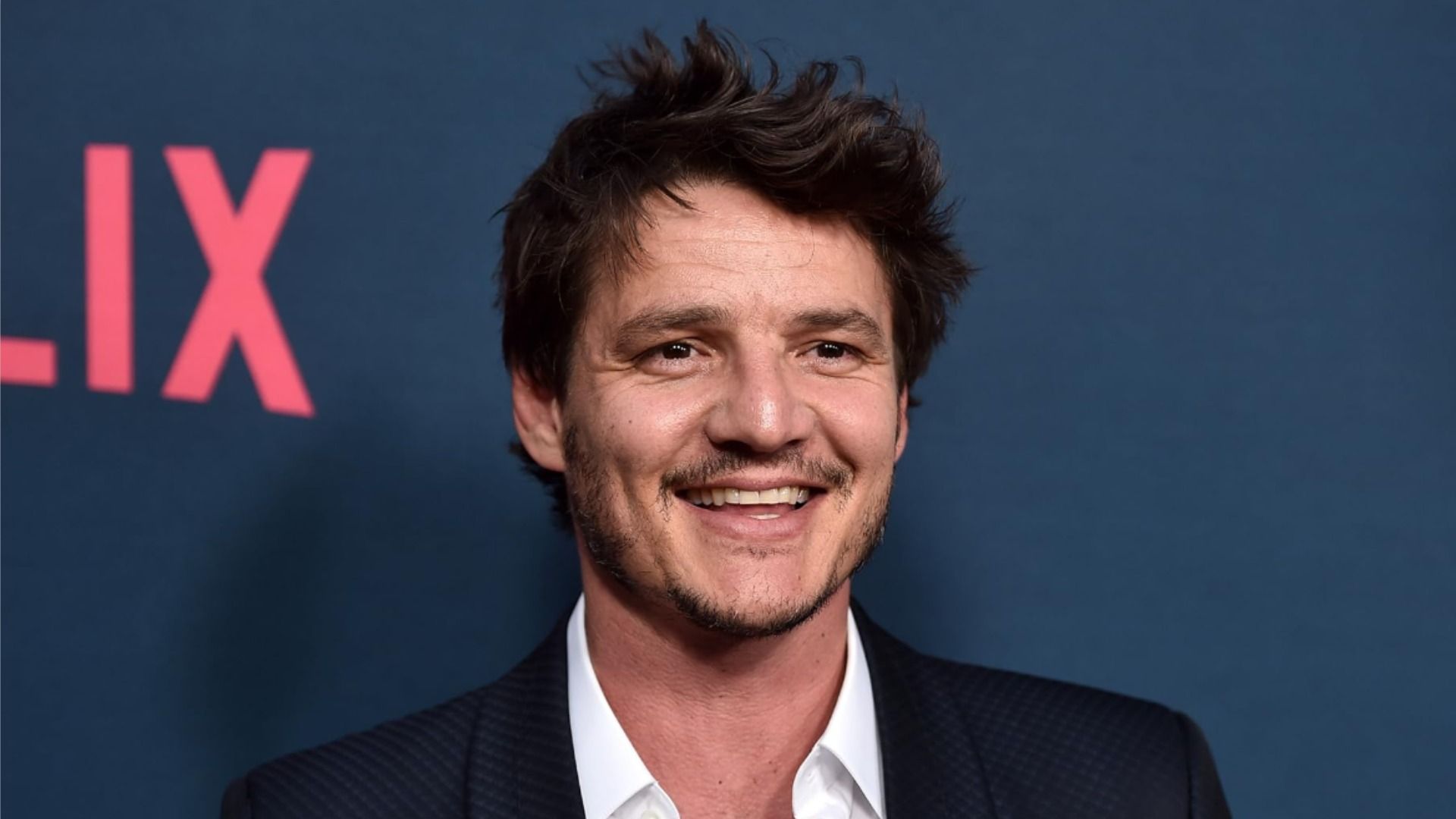 L'attore Pedro Pascal in giacca nera sorride su sfondo blu
