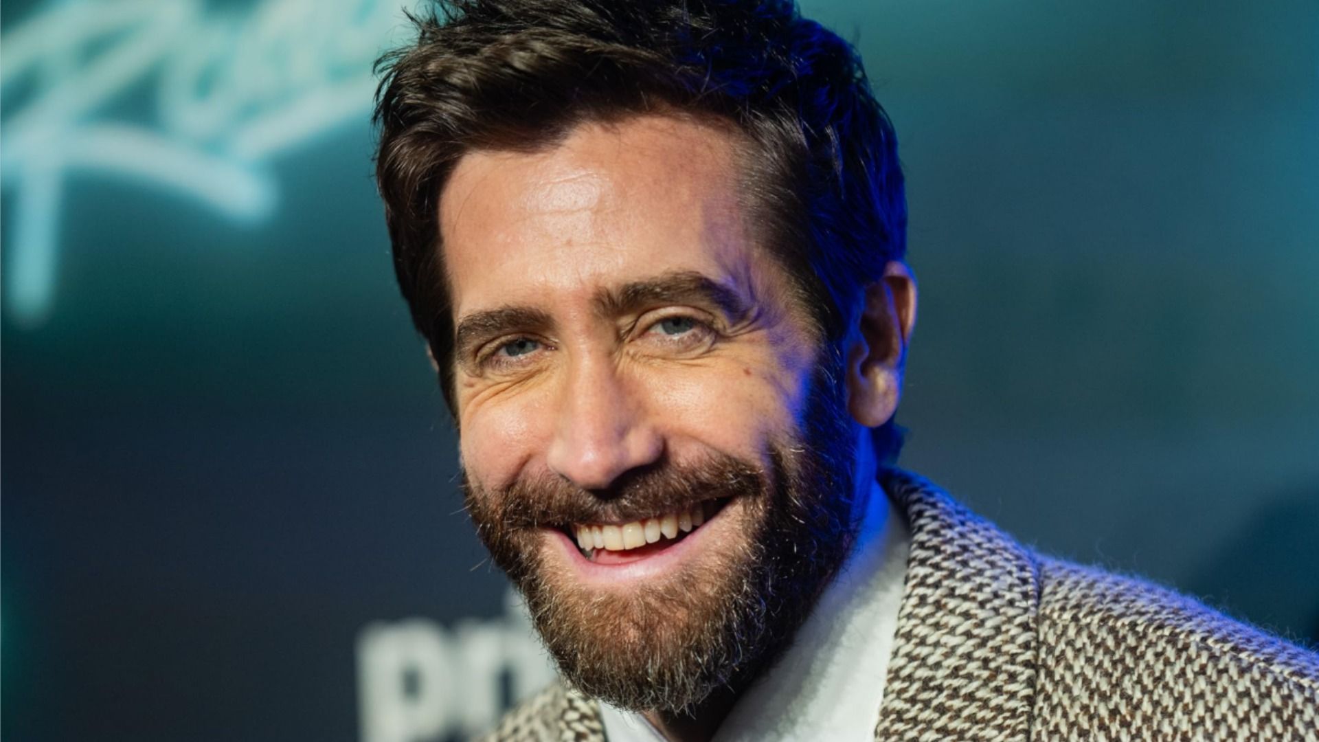 L'attore Jake Gyllenhaal in un blazer grigio sorride su uno sfondo blu