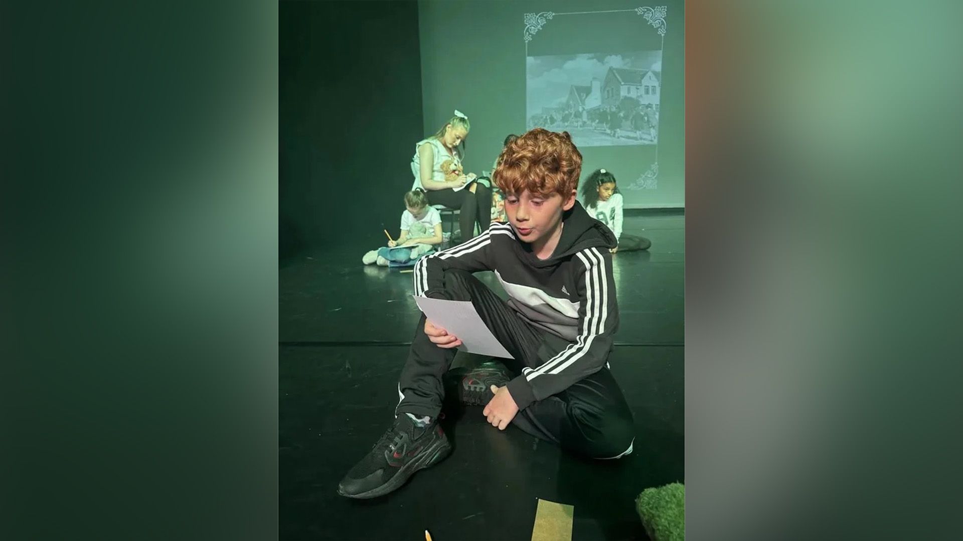 Alastair Stout in tuta seduto sul palco di un teatro mentre legge da un foglio