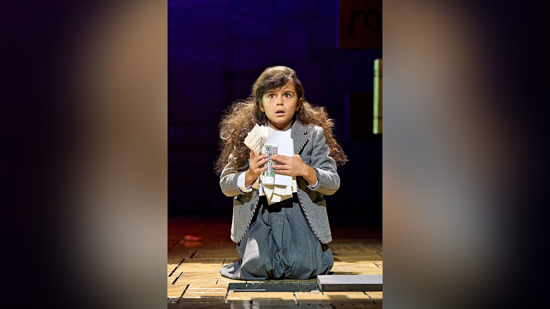 Arabella Stanton in un vestito grigio inginocchiata su un palcoscenico teatrale, tiene in mano dei fogli accartocciati