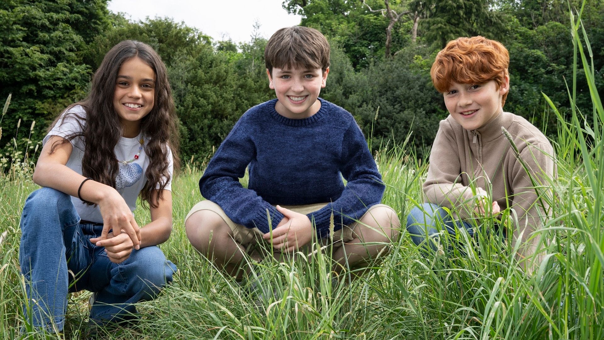 I giovani attori Arabella Stanton, Dominic McLaughlin e Alastair Stout accovacciati nell'erba che sorridono