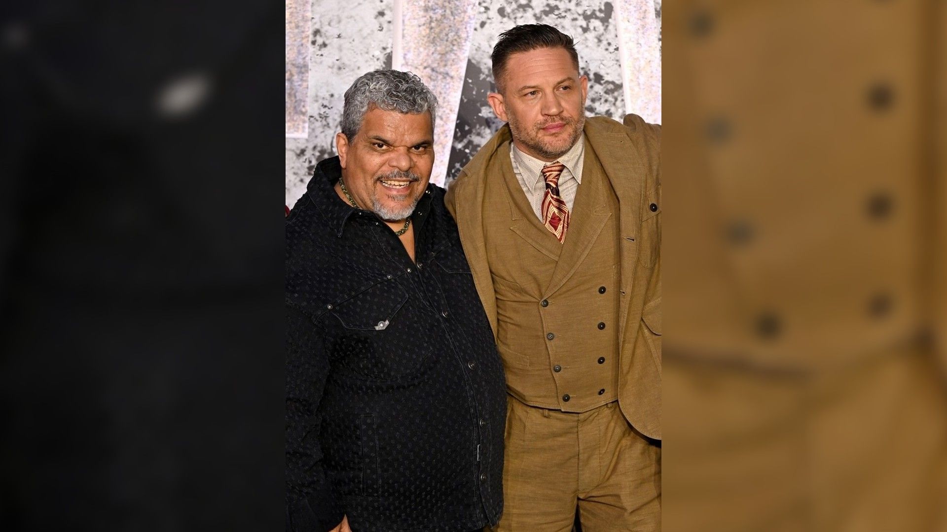 Luis Guzmán e Tom Hardy posano sul red carpet: uno in camicia scura, l'altro in completo marrone con cravatta