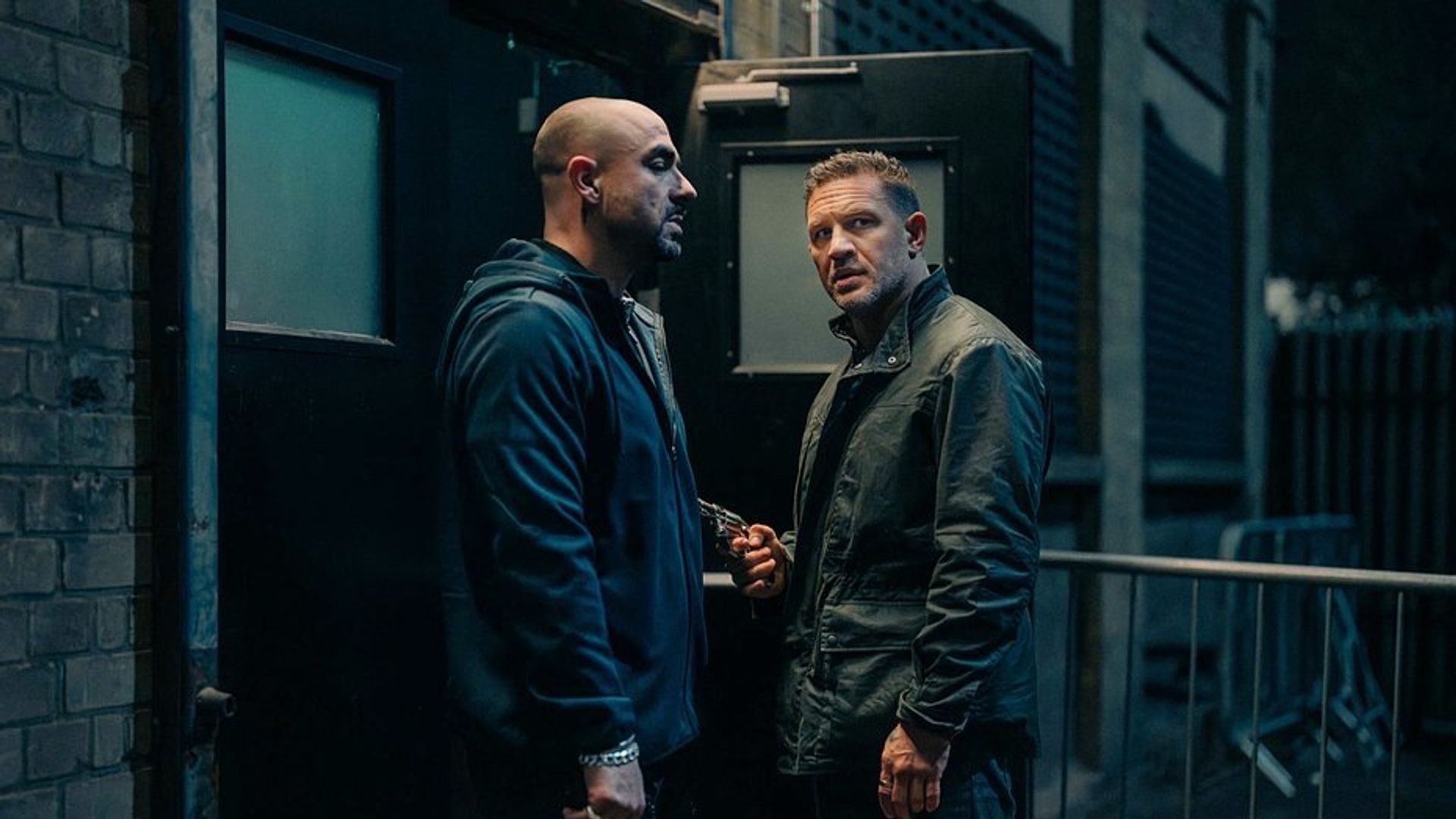 Tom Hardy e un altro uomo in abiti scuri stanno parlando vicino a un'uscita nera di notte, uno di loro tiene un'arma