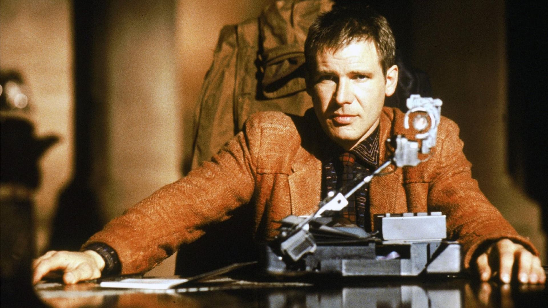 L'attore Harrison Ford nei panni di Rick Deckard seduto a un tavolo con una giacca marrone