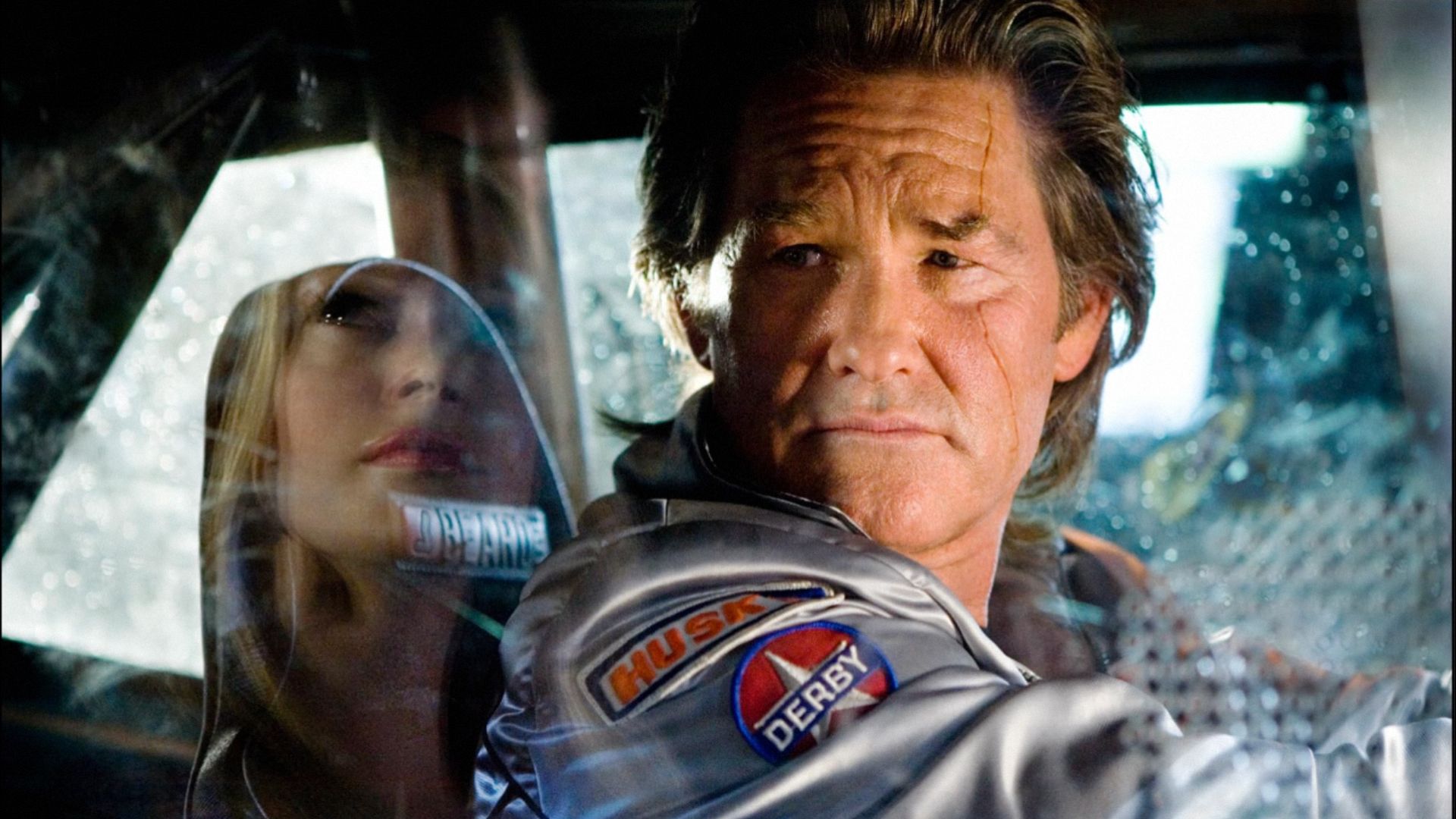 L'attore Kurt Russell in giacca argentata seduto al volante di un'auto e guarda di lato