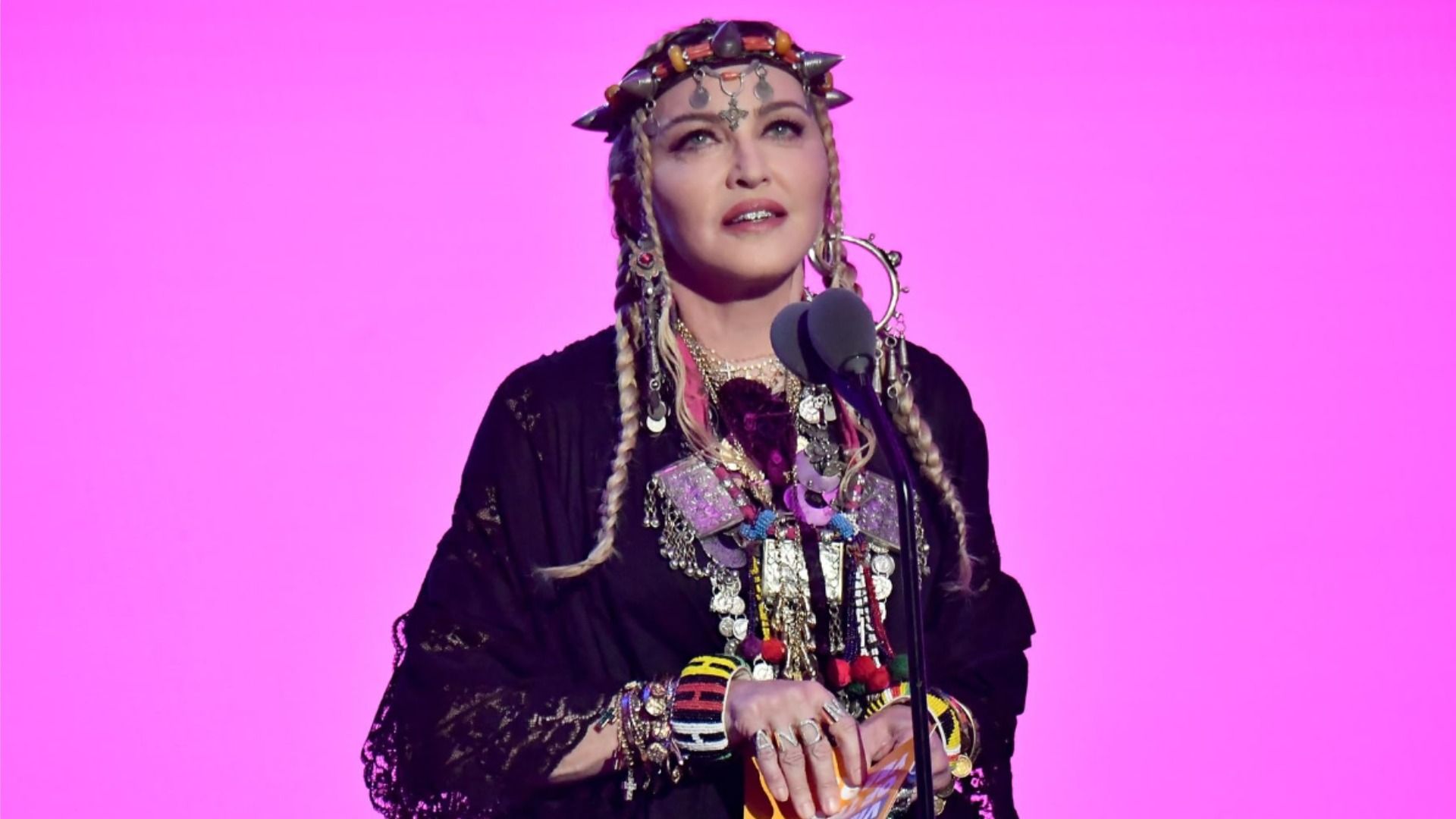 La cantante Madonna in un vestito nero su uno sfondo rosa guarda verso l'alto