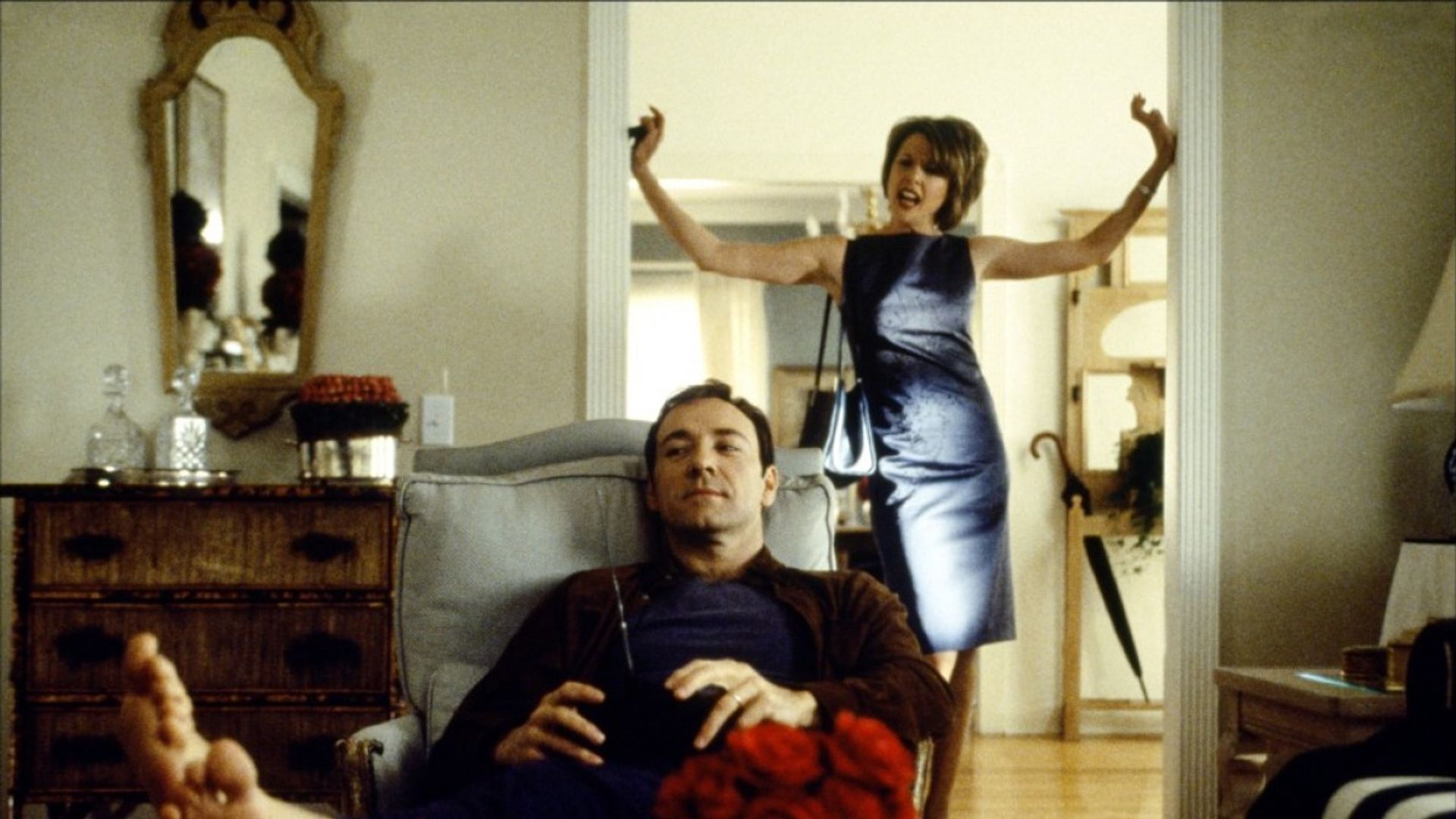 Kevin Spacey seduto rilassato su una poltrona con un telecomando mentre una donna in abito da sera dietro di lui gesticola emotivamente sulla soglia
