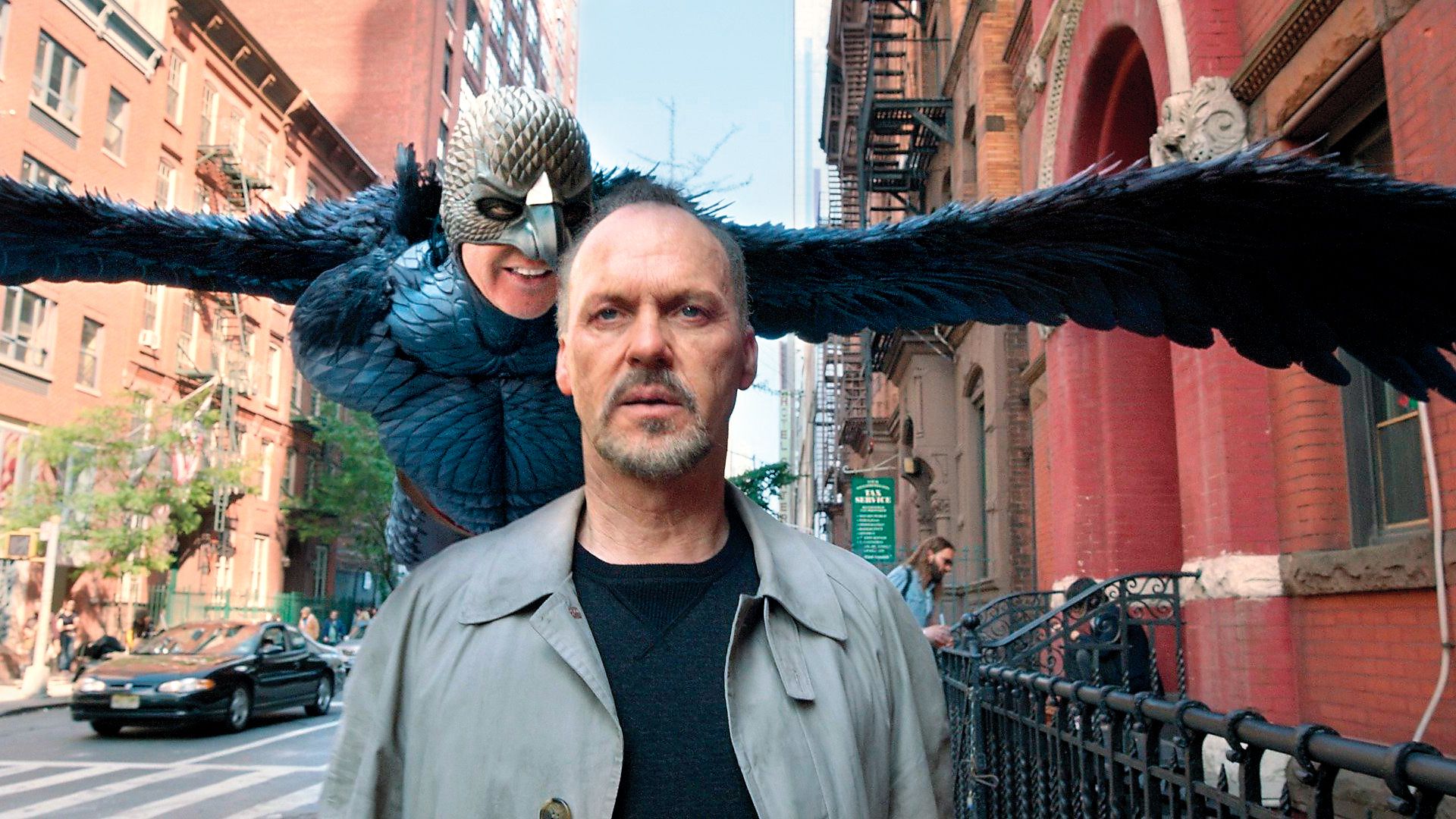 Michael Keaton cammina per strada mentre dietro di lui aleggia un personaggio fantastico con ali e maschera da uccello