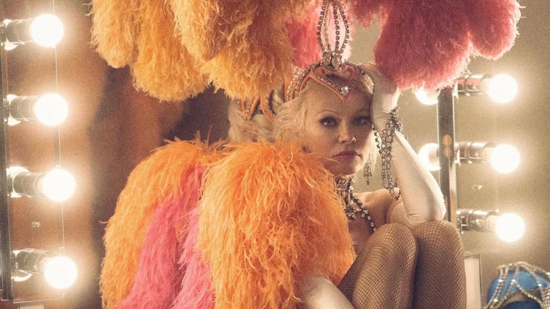 Pamela Anderson in un costume da spettacolo scintillante con piume e decorazioni siede davanti allo specchio del camerino, appoggiando pensierosa la testa sulla mano
