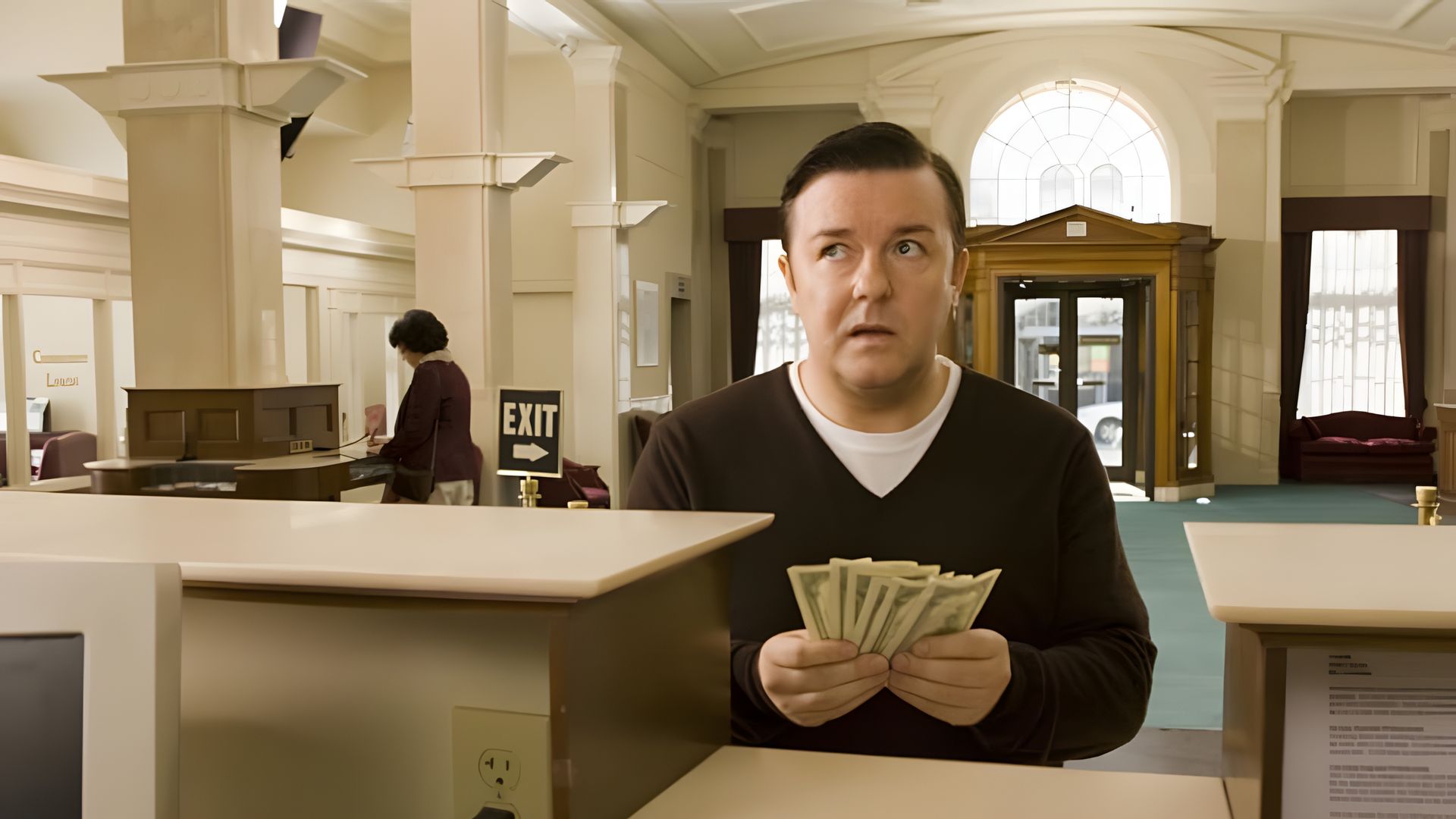 Ricky Gervais con un'espressione sorpresa tiene una pila di soldi in una banca, in piedi al bancone sullo sfondo di un'uscita e finestre alte