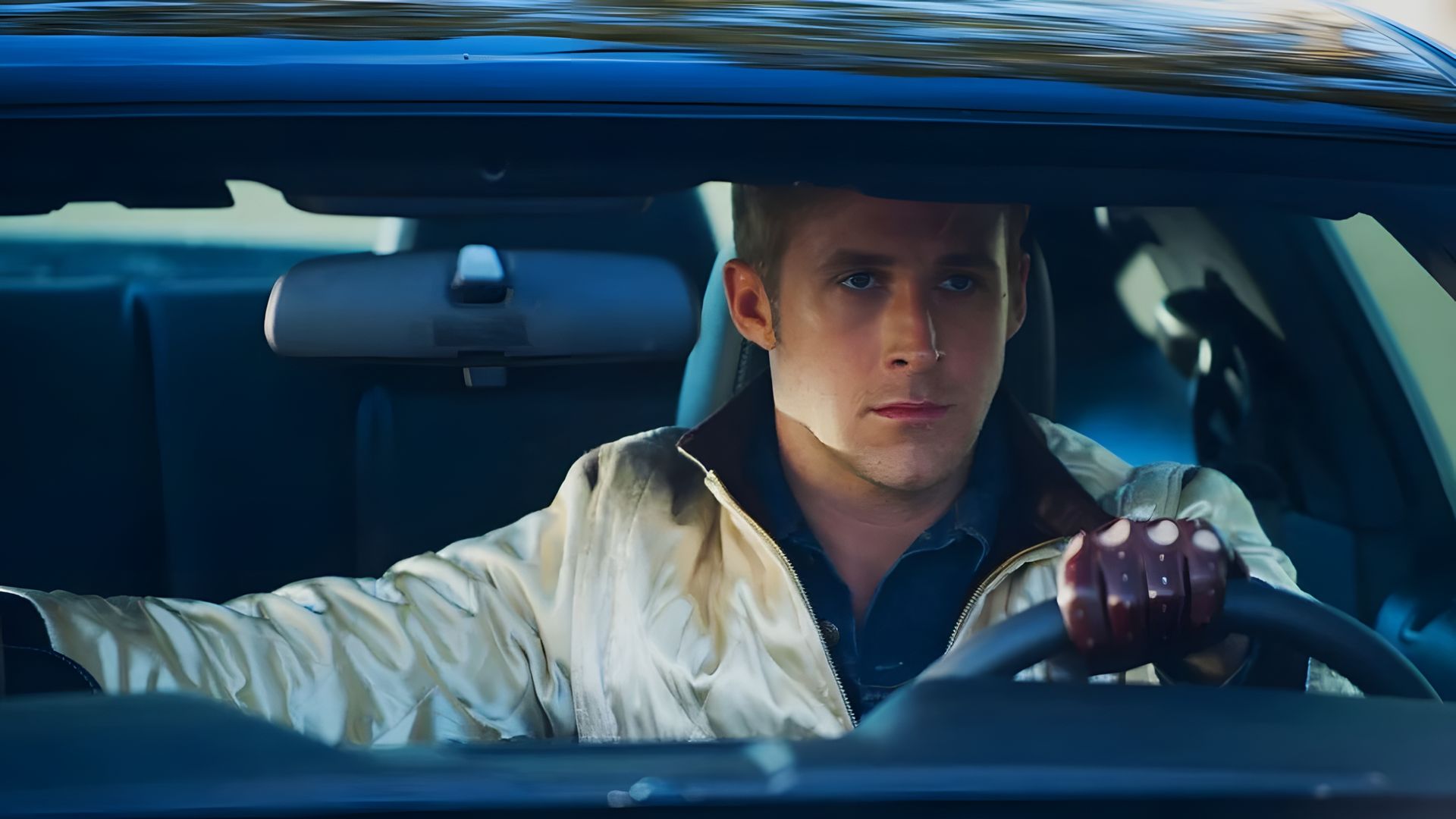 Ryan Gosling in giacca di raso e guanti di pelle seduto al volante di un'auto, guarda avanti con espressione concentrata