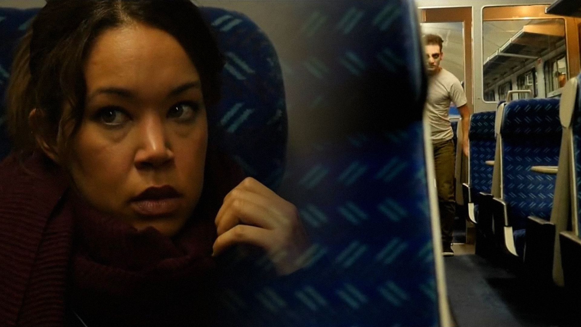 Una ragazza con una sciarpa bordeaux seduta in una carrozza del treno mentre un uomo in maglietta bianca cammina tra i sedili nelle vicinanze