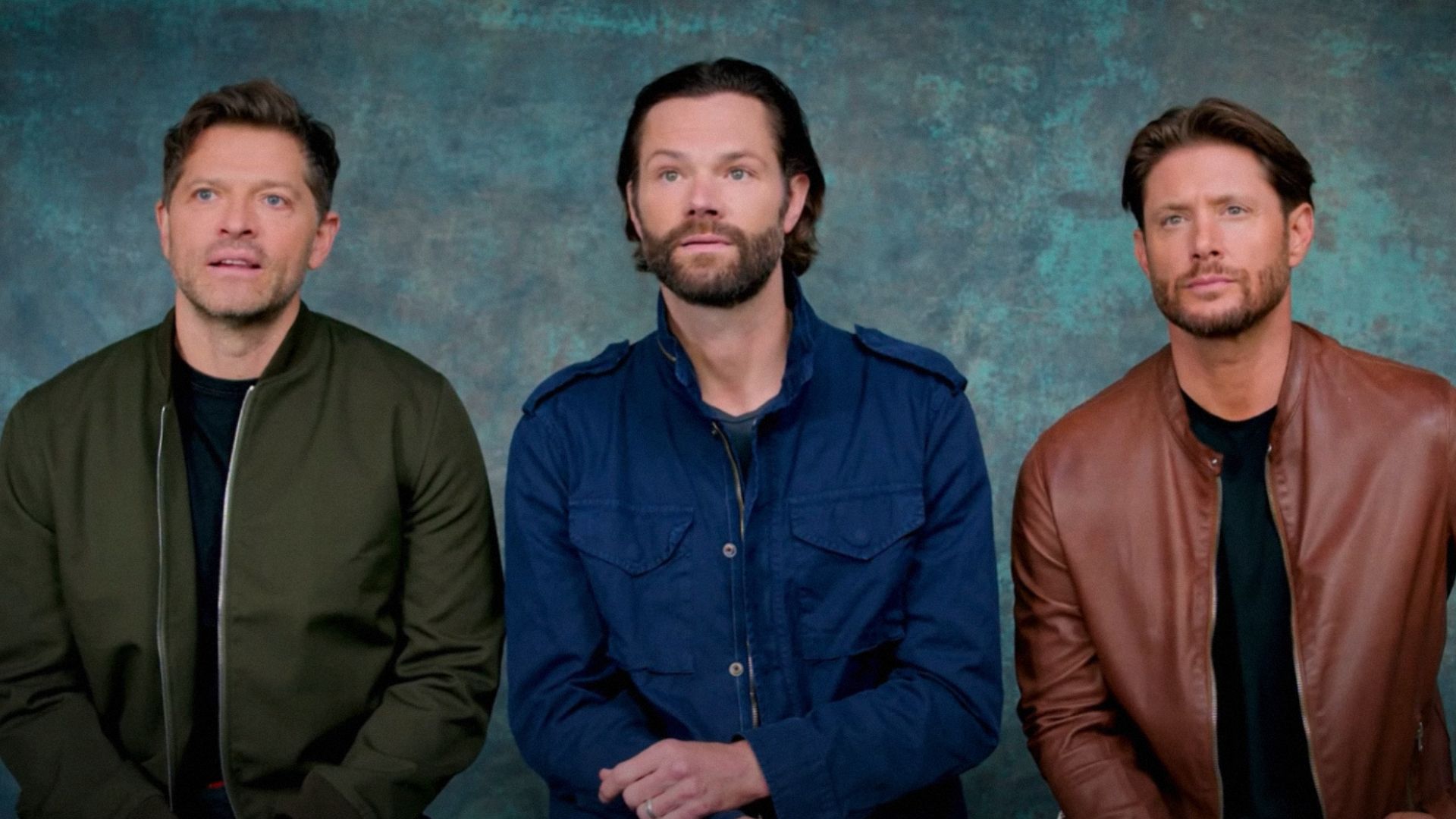 Gli attori Misha Collins, Jared Padalecki e Jensen Ackles seduti contro uno sfondo blu indossando giacche colorate