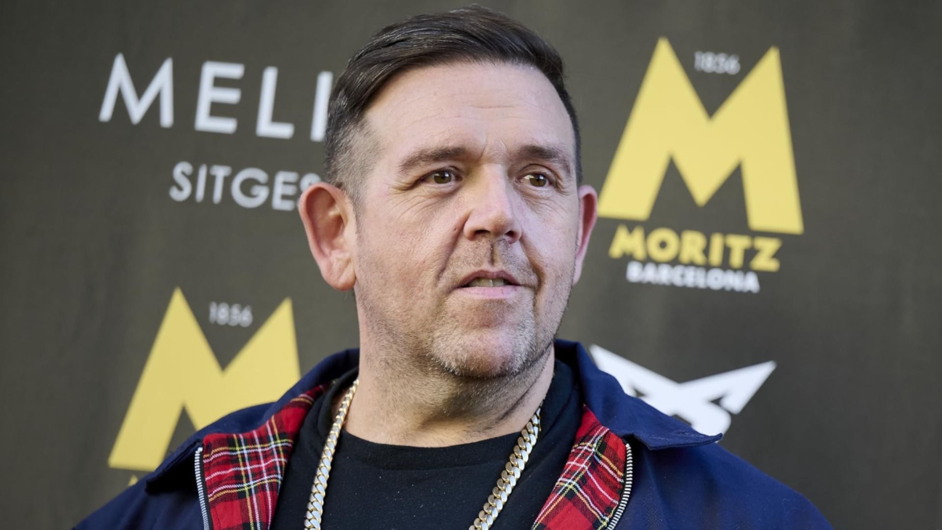 L'attore Nick Frost in una giacca blu con una catena d'oro al collo sorride davanti a uno sfondo espositivo