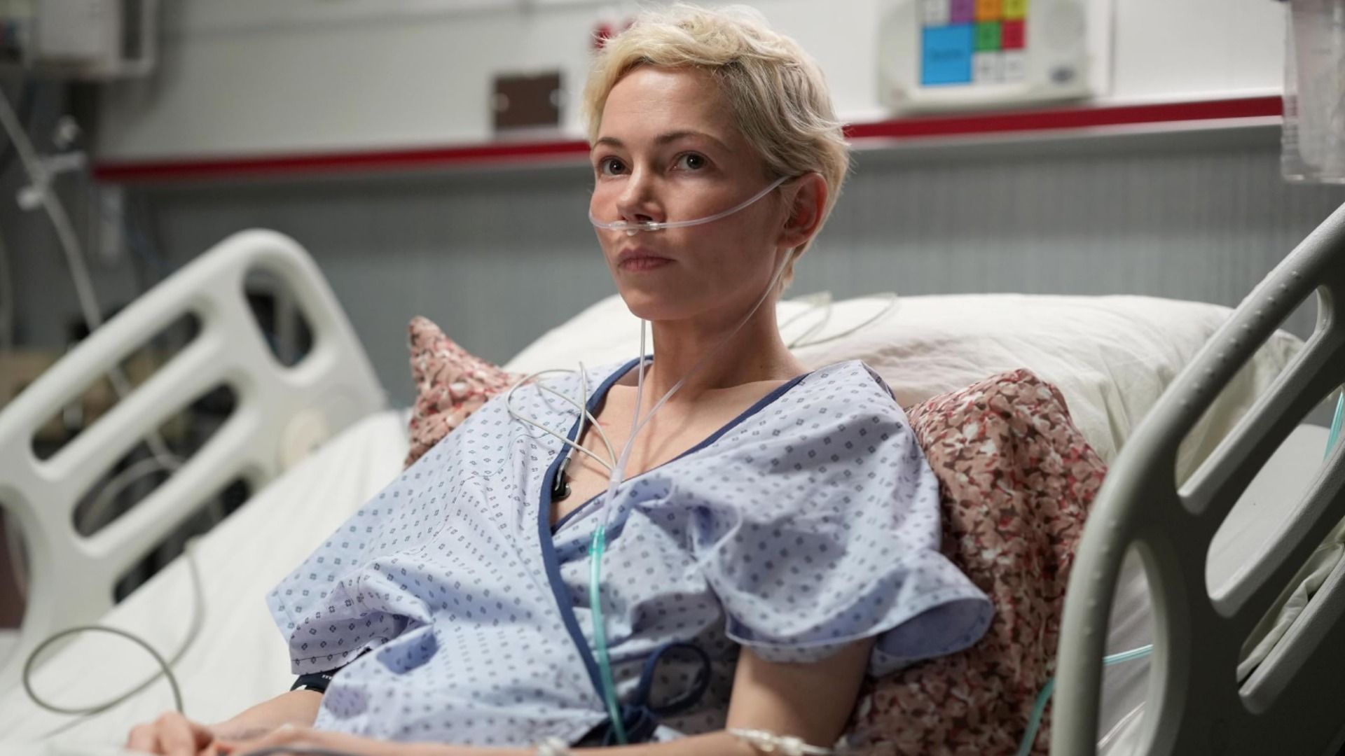 L'attrice Michelle Williams in abiti da paziente ospedaliera giace su un letto con un tubo nel naso