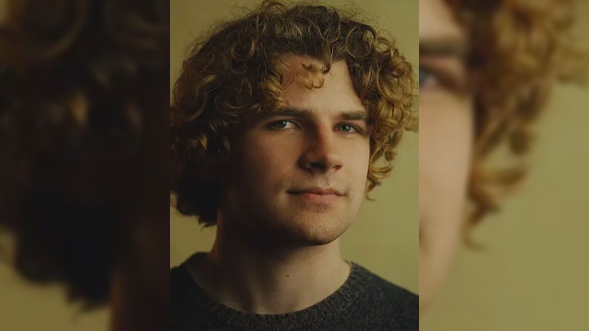 Un giovane uomo dai capelli ricci in un maglione nero sta in piedi contro uno sfondo giallo