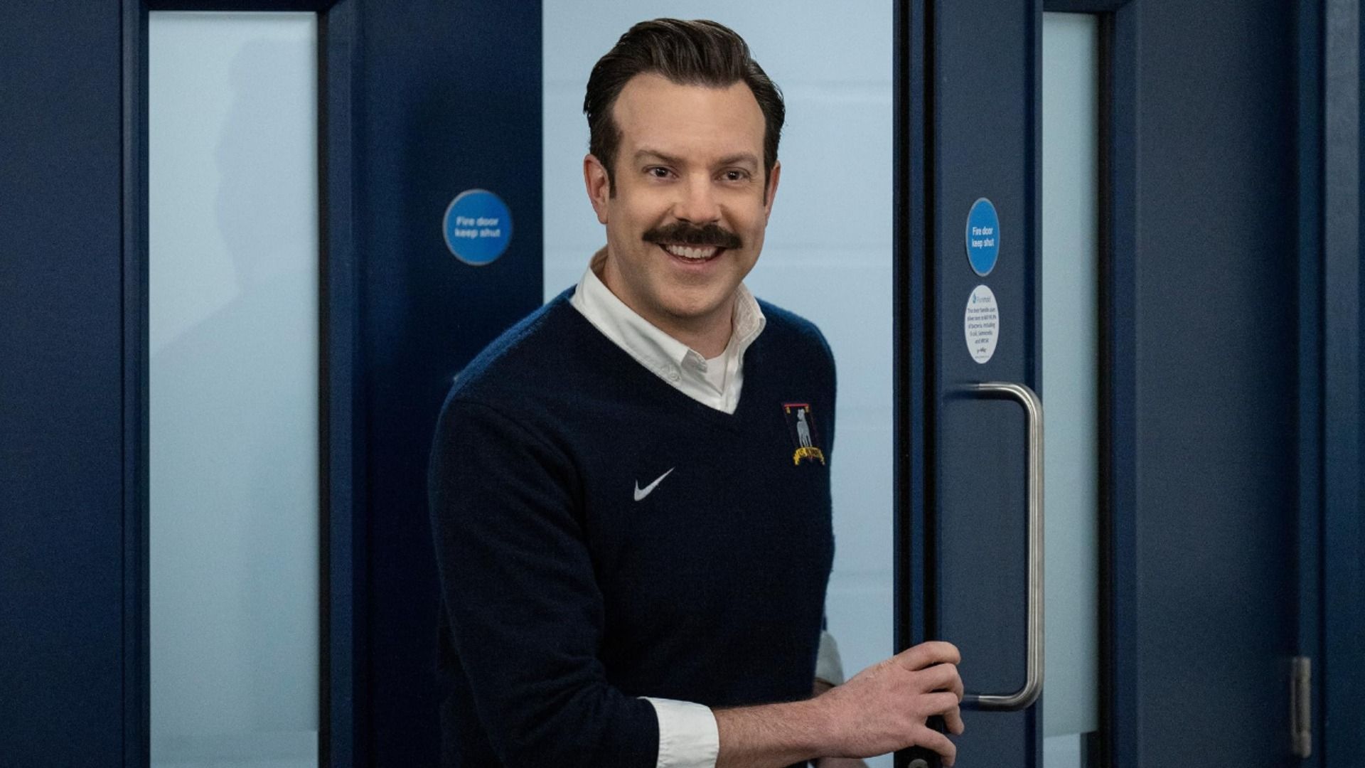 L'attore Jason Sudeikis con un maglione blu e i baffi in piedi vicino a una porta che sorride