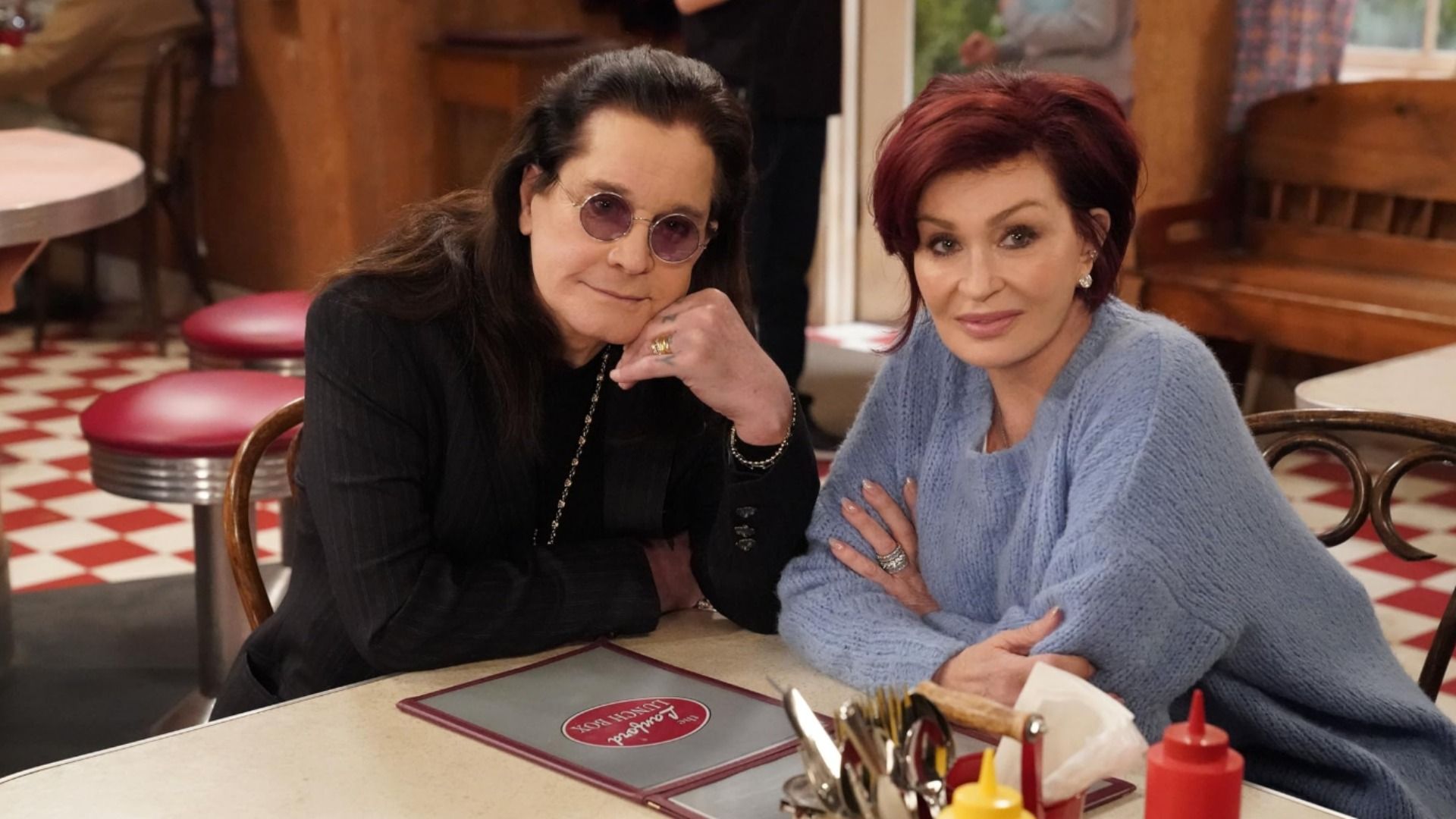 Il cantante Ozzy Osbourne con occhiali e giacca nera seduto a un tavolo con la moglie