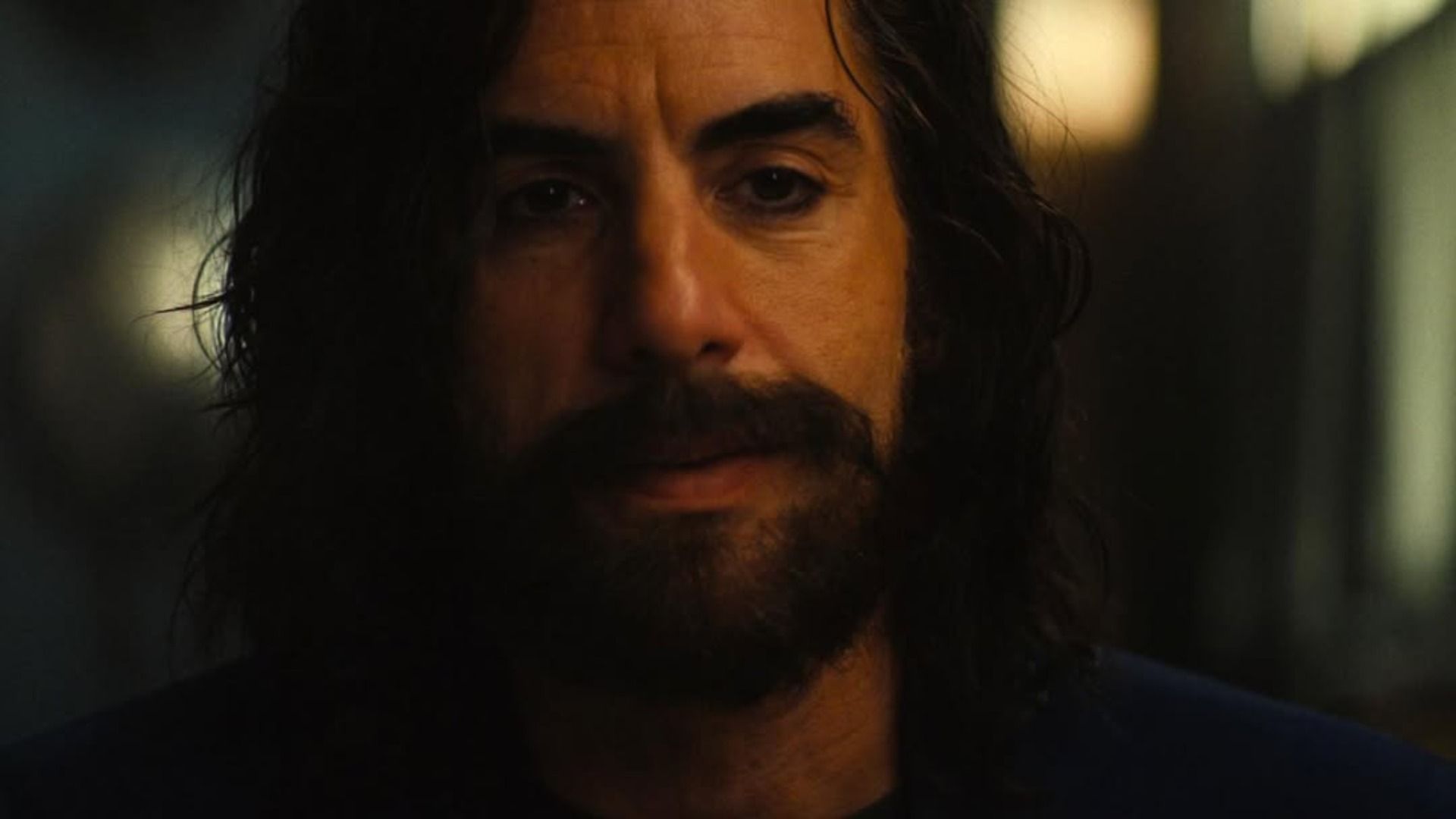 L'attore Sacha Baron Cohen con barba e capelli lunghi in piedi contro uno sfondo sfocato