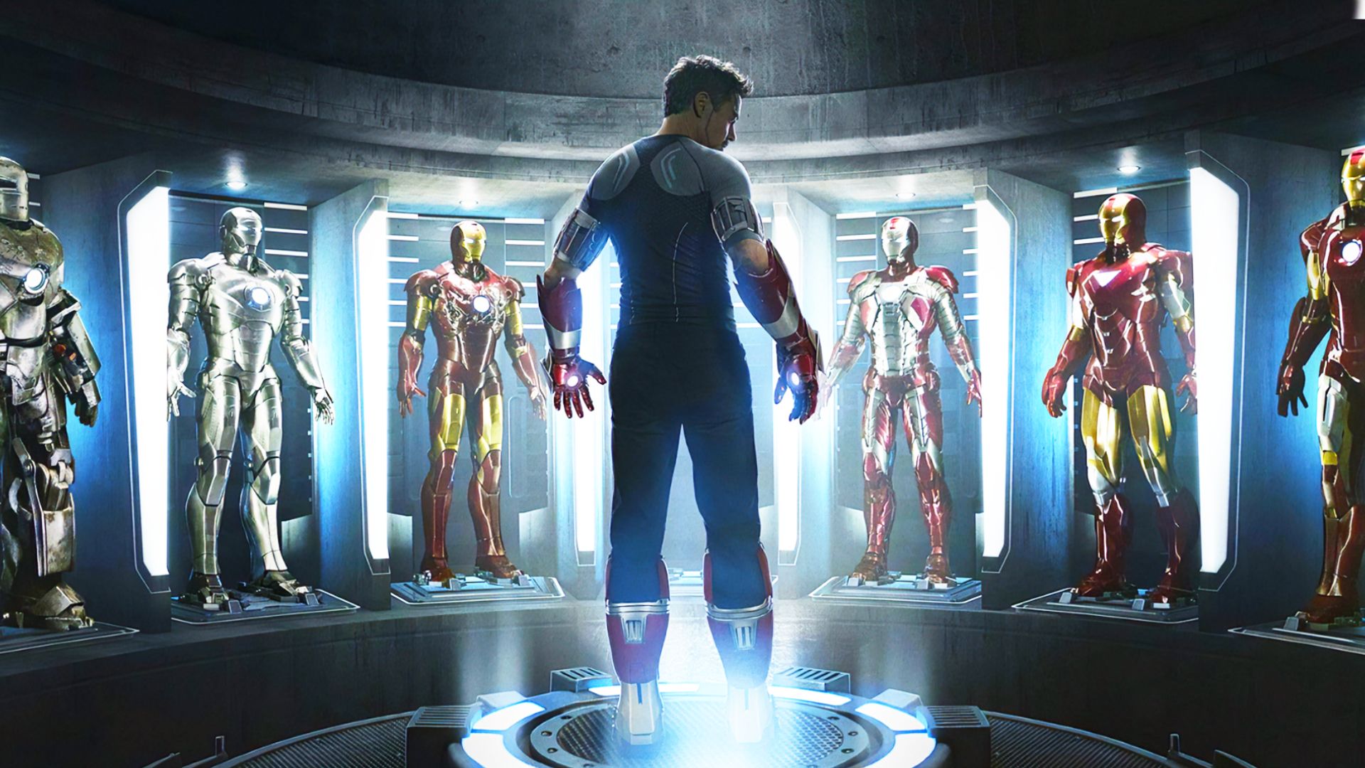 L'attore Robert Downey Jr. in abiti grigi e neri sta in piedi circondato dalle armature di Iron Man