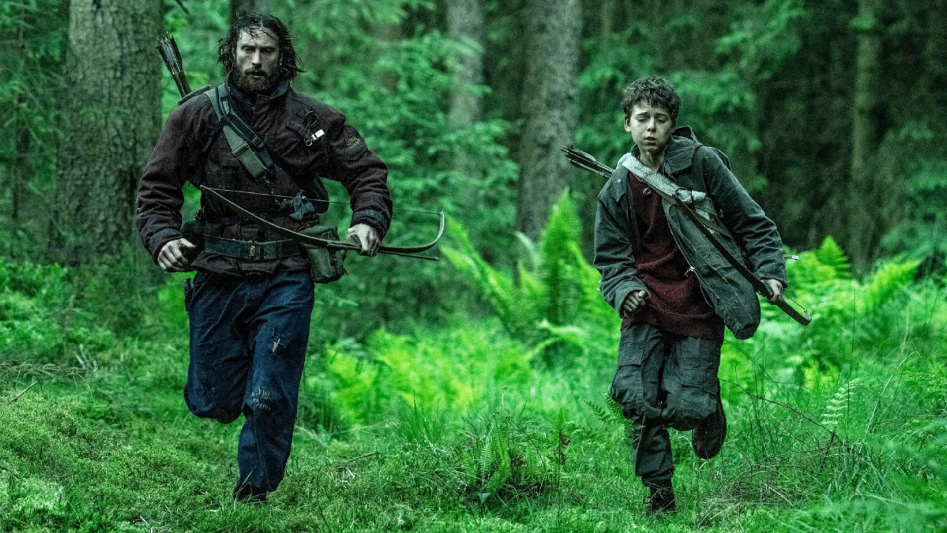 L'attore Aaron Taylor-Johnson in abiti sporchi corre attraverso la foresta insieme a un bambino con l'arco