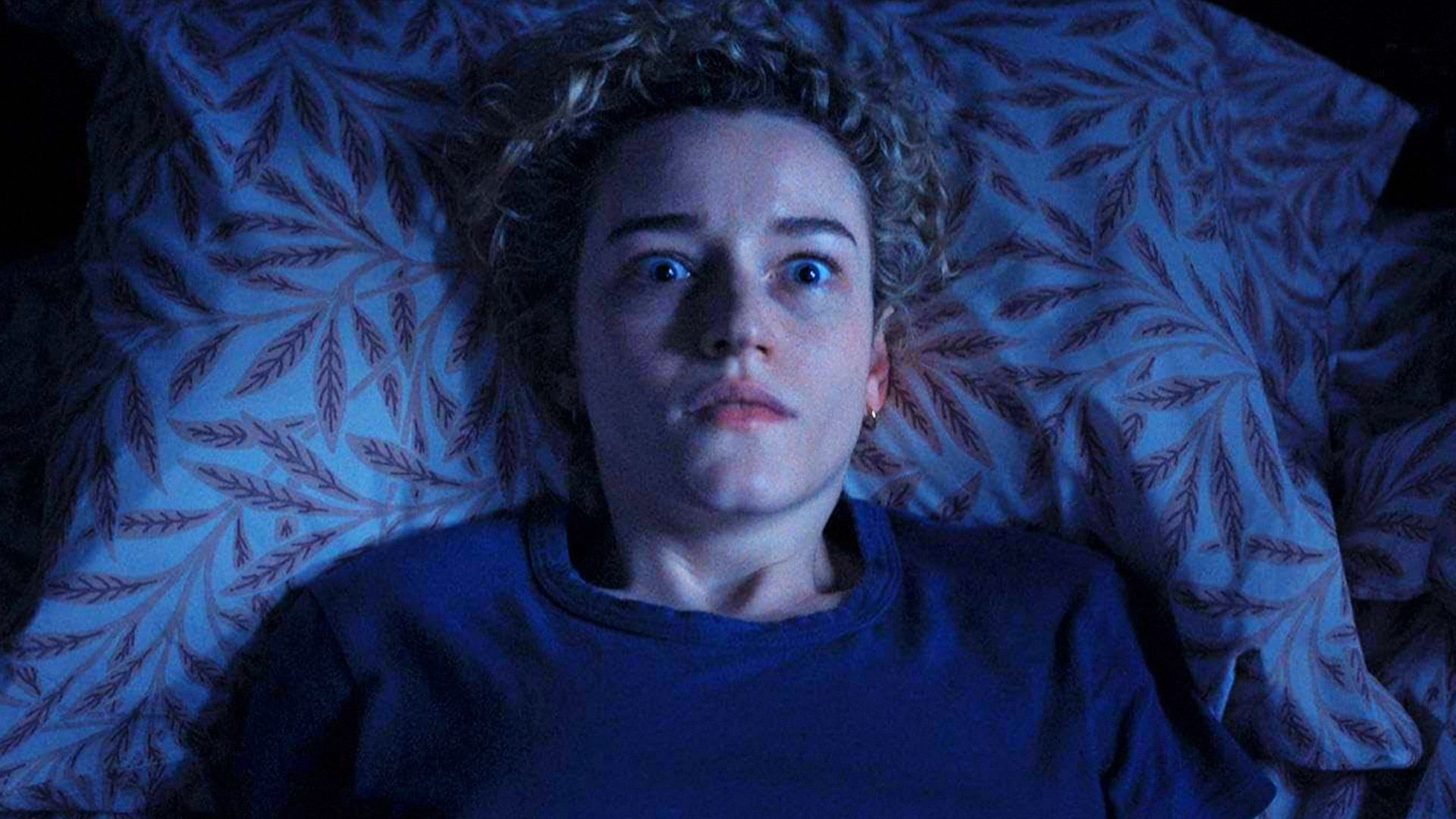 L'attrice Julia Garner in un maglione blu giace su un letto, spaventata da qualcosa