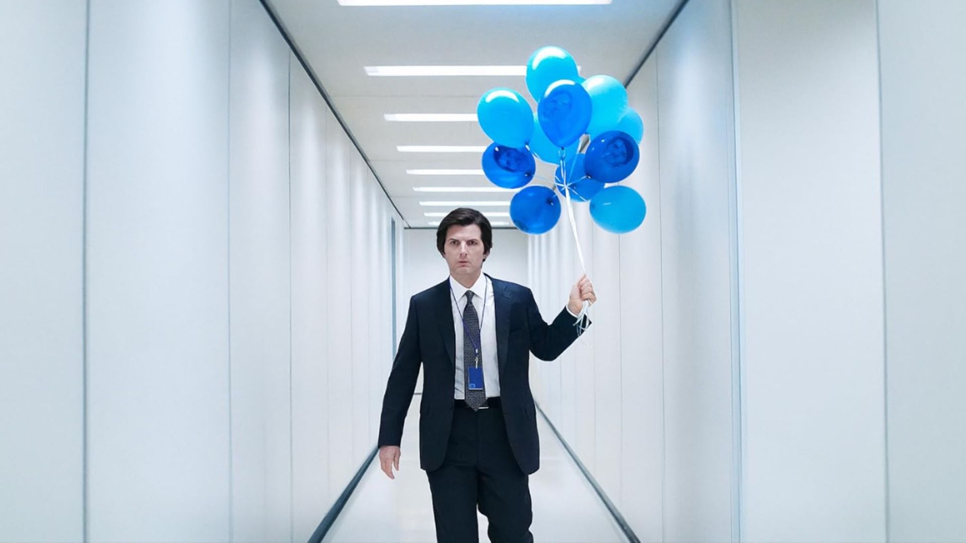 L'attore Adam Scott in un completo classico cammina lungo un corridoio bianco tenendo palloncini blu