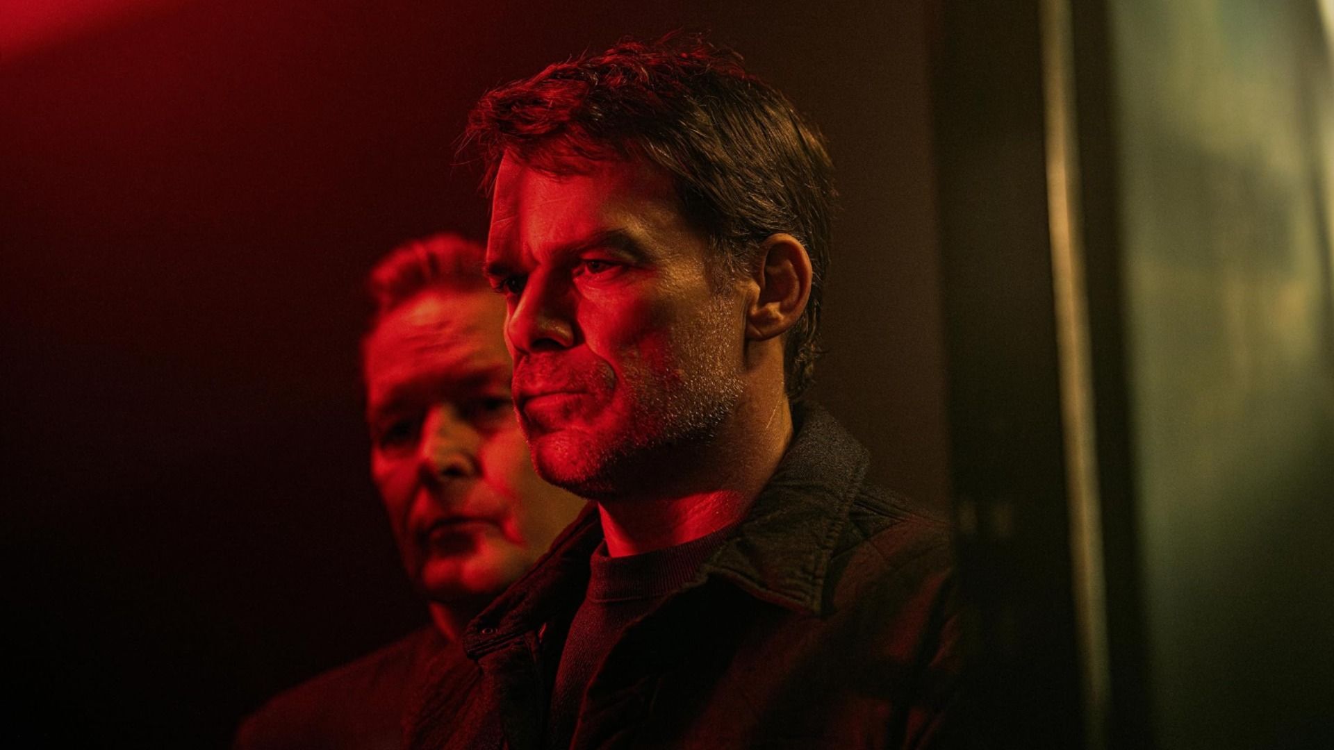 L'attore Michael C. Hall sta in profilo sotto una luce rossa con parte del volto di un uomo visibile dietro di lui