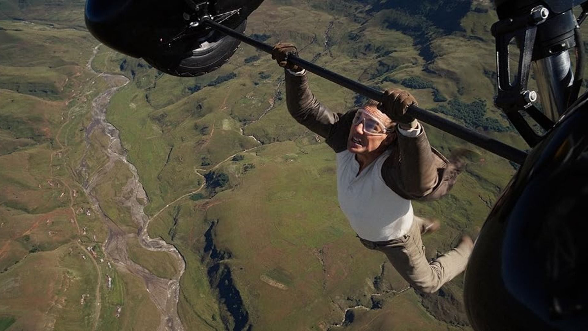 L'attore Tom Cruise in giacca marrone si aggrappa alle ruote di un aereo in volo