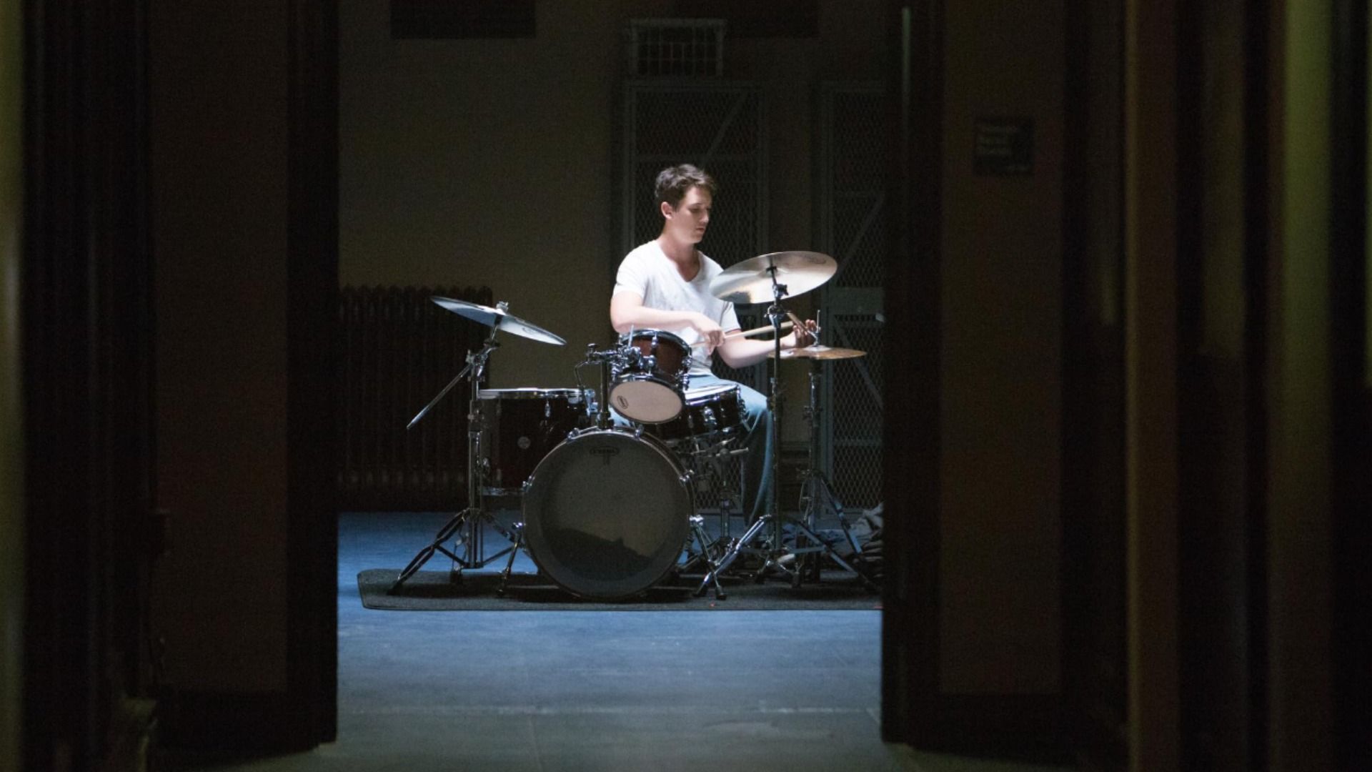 Illuminazione soffusa, l'attore Miles Teller suona la batteria in una maglietta bianca
