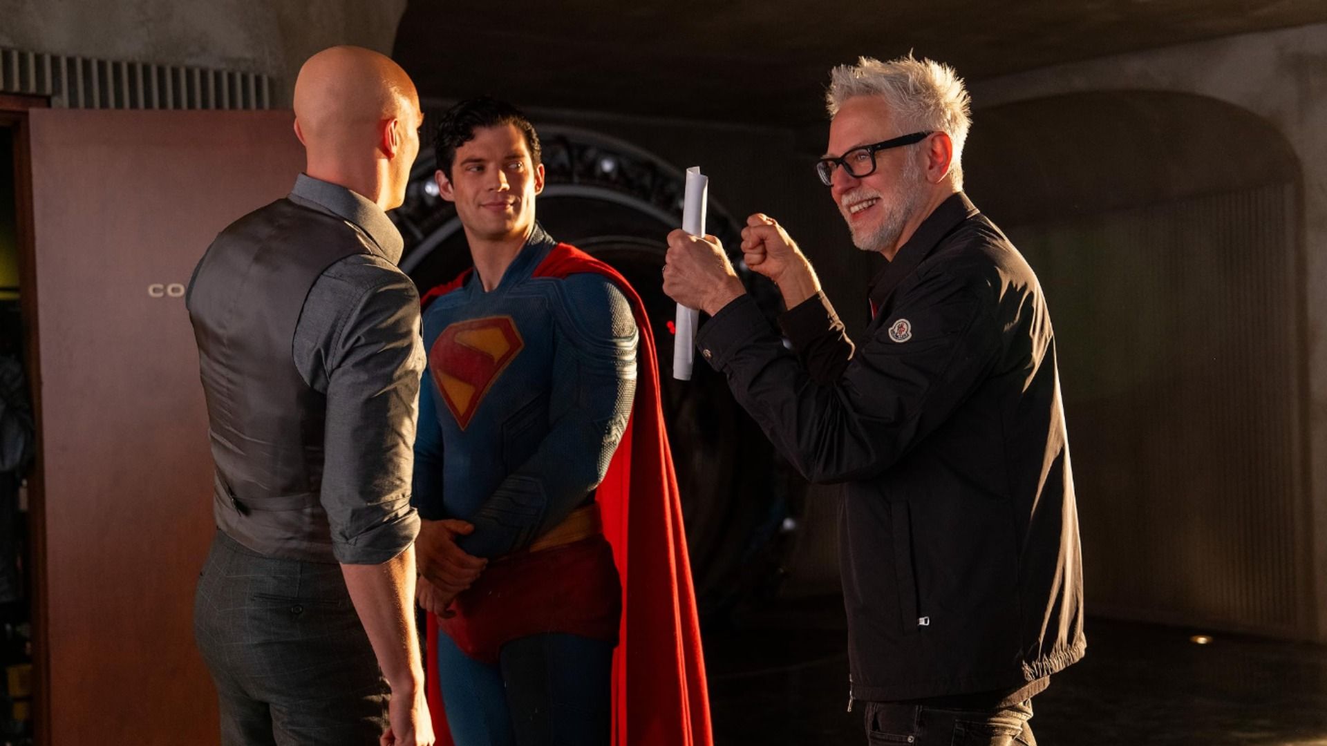 Il regista James Gunn con carte in mano parla con l'attore David Corenswet in costume da Superman e con Nicholas Hoult che è calvo