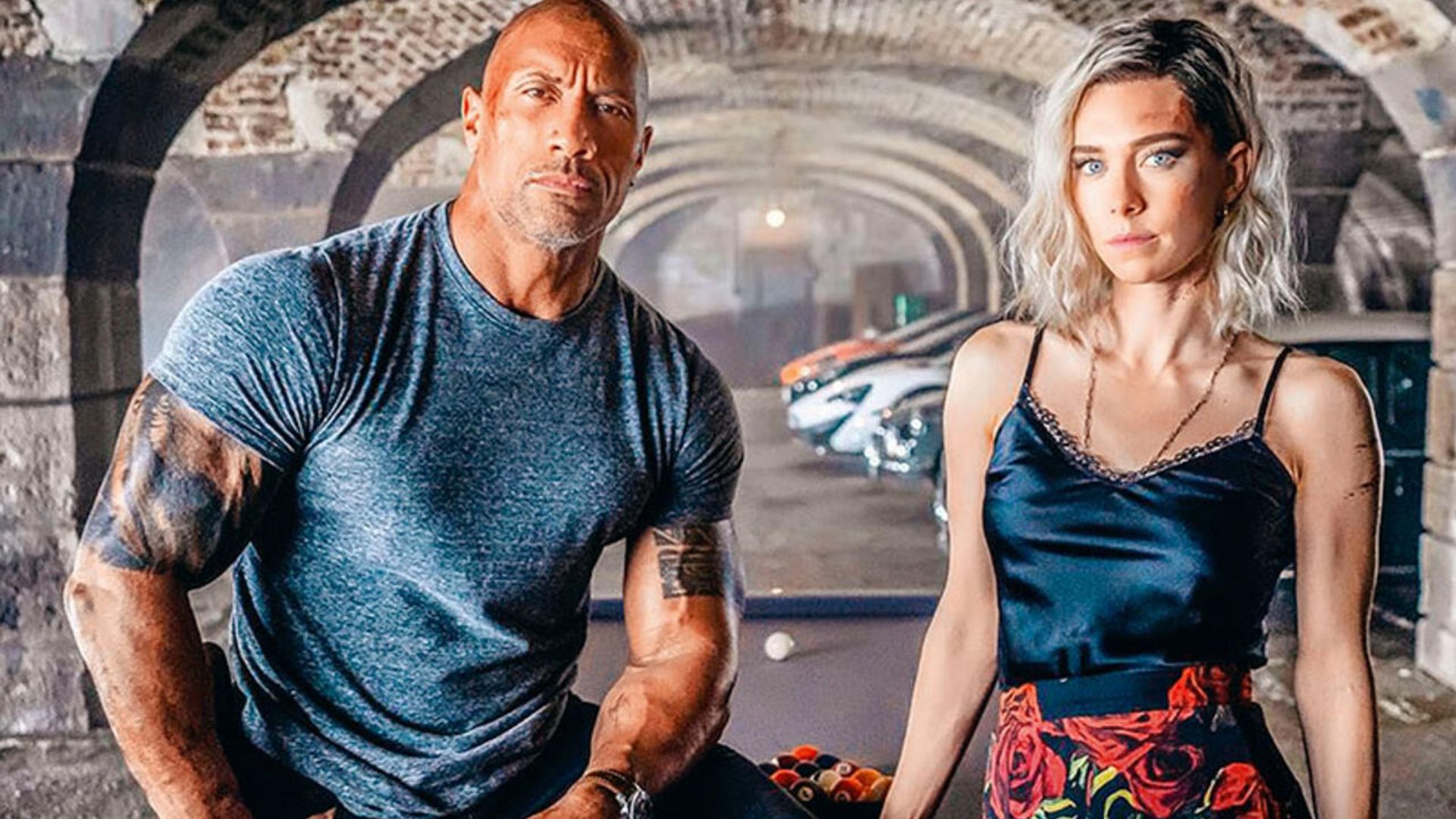 Dwayne Johnson in una maglietta aderente e Vanessa Kirby in canottiera e gonna fantasia posano davanti a un fondale di archi e auto sportive
