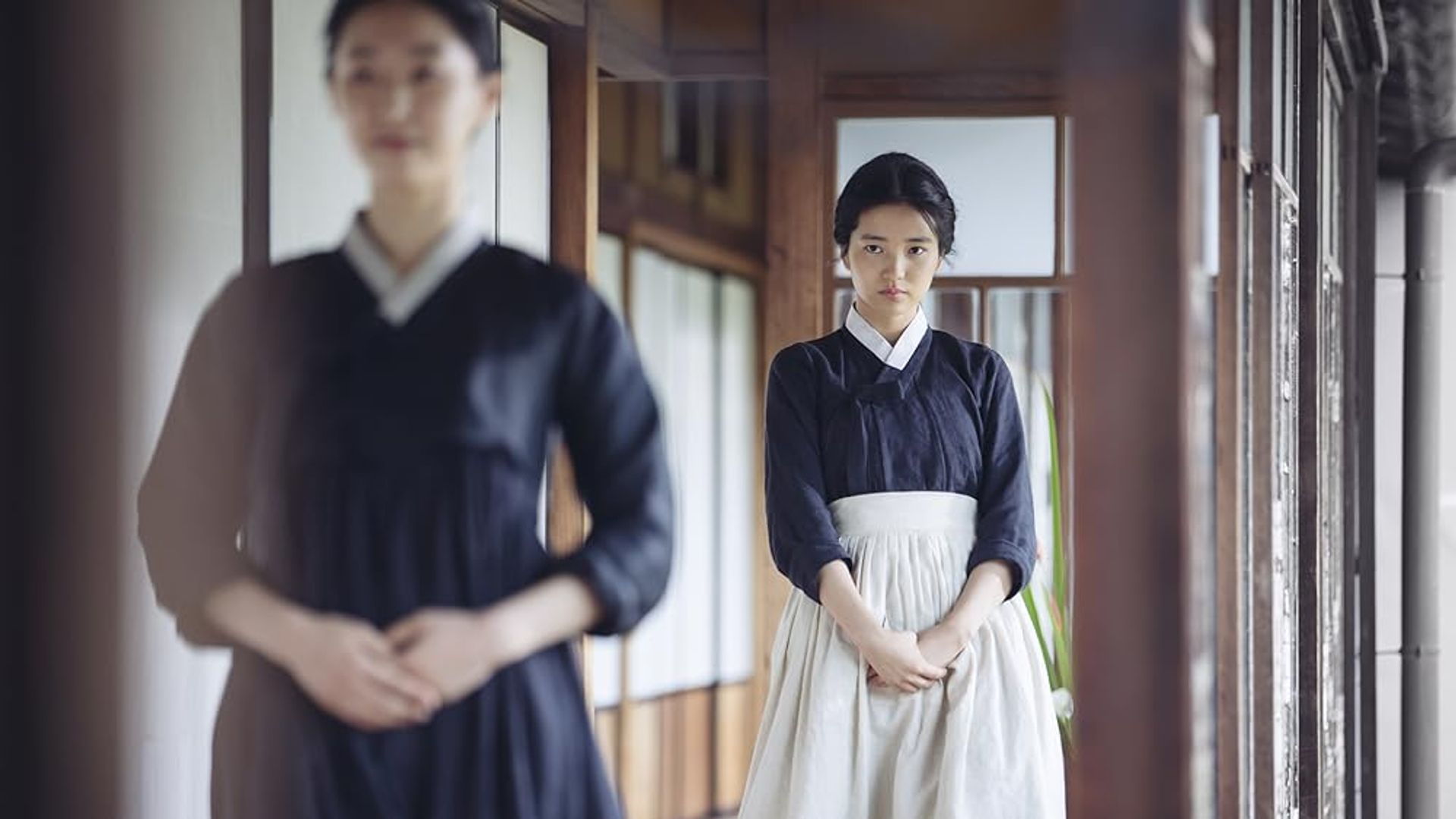 Una ragazza in hanbok nero con grembiule bianco sta in piedi nel corridoio, guardando avanti con espressione seria