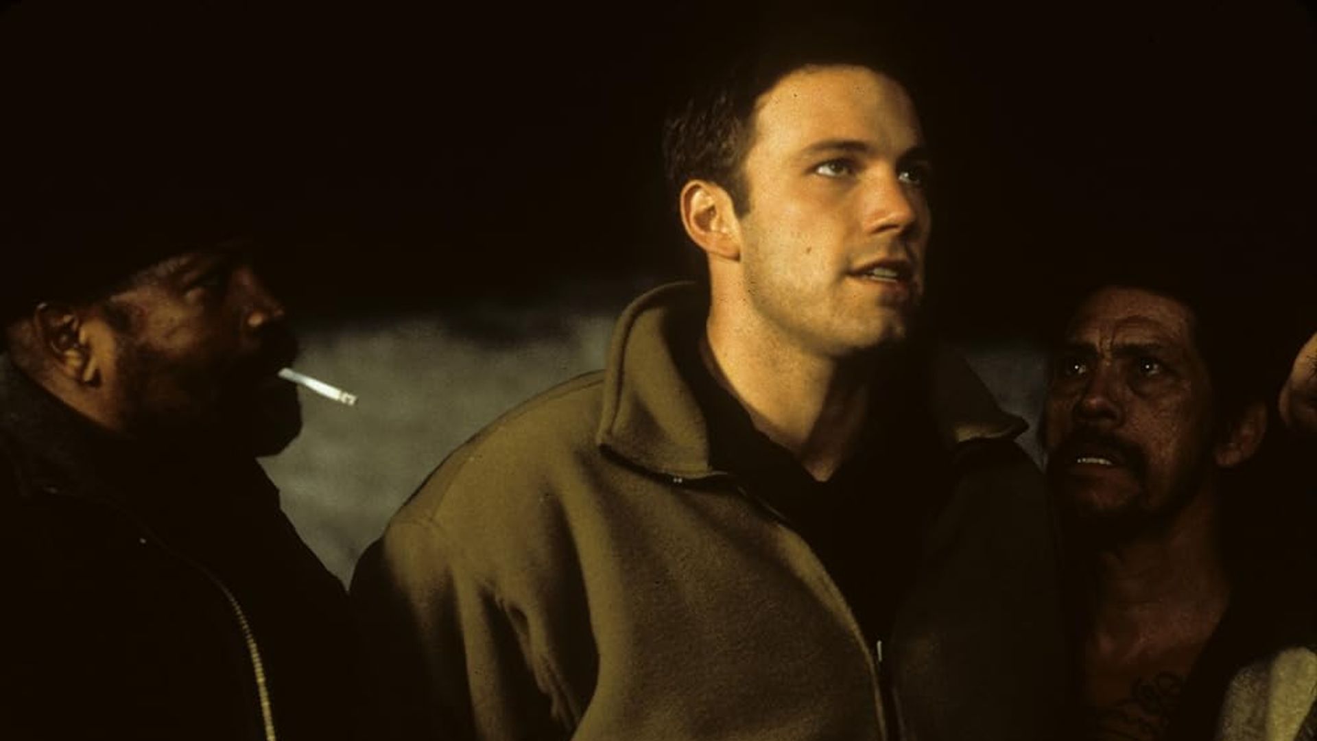Ben Affleck con una giacca beige sta tra due persone, una delle quali sta fumando, in un ambiente poco illuminato