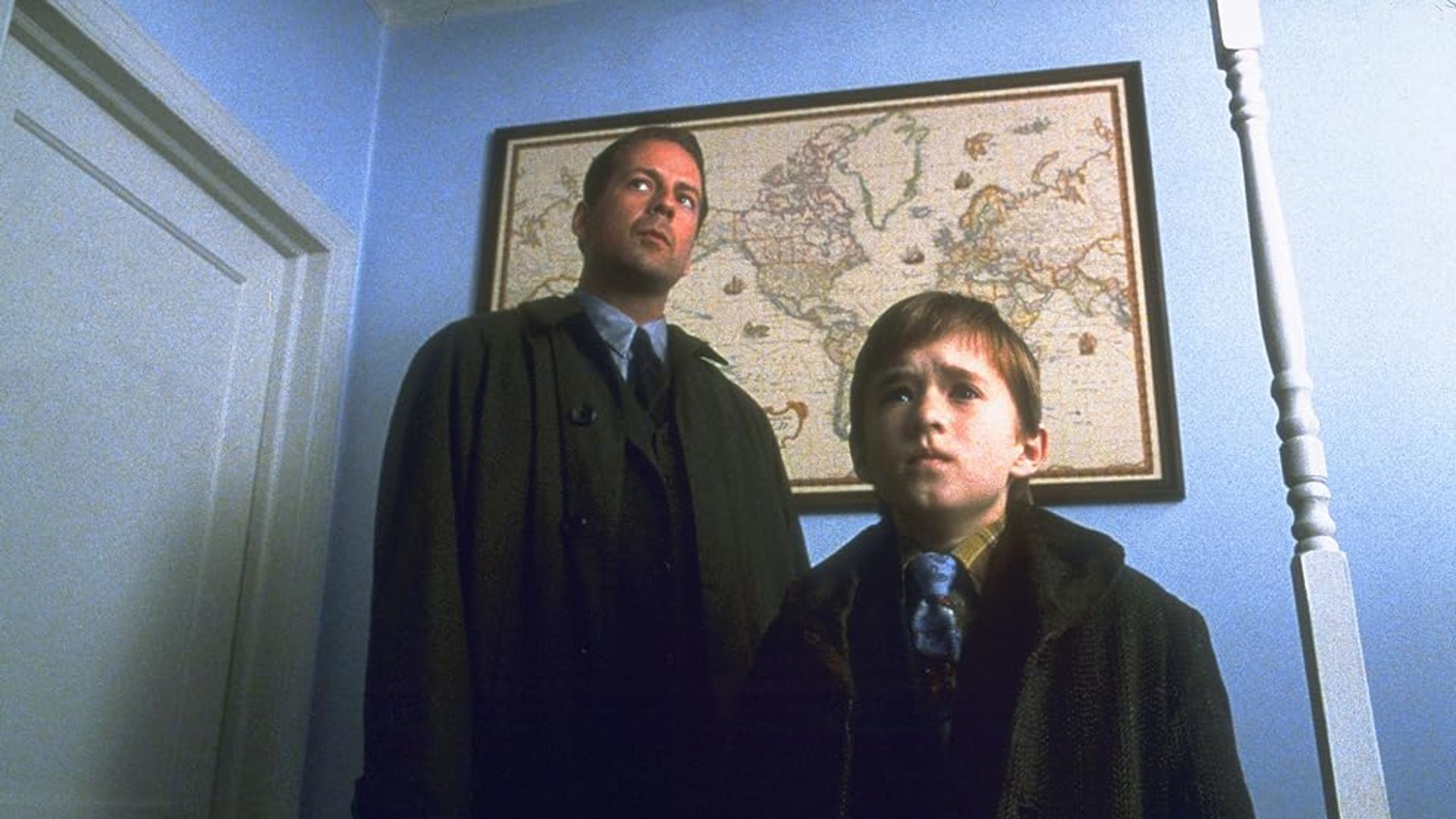 Bruce Willis e Haley Joel Osment in piedi in un corridoio con una mappa del mondo appesa al muro dietro di loro
