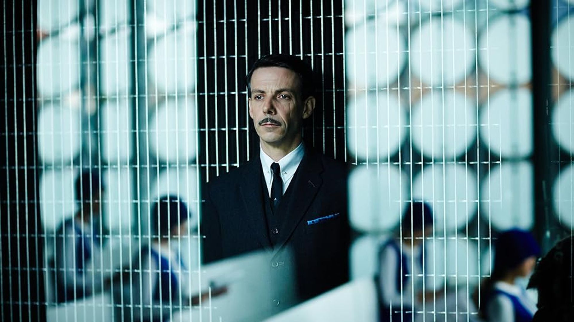 Ethan Hawke in completo con i baffi dietro una parete a rete con persone in uniforme che lavorano alle scrivanie sullo sfondo