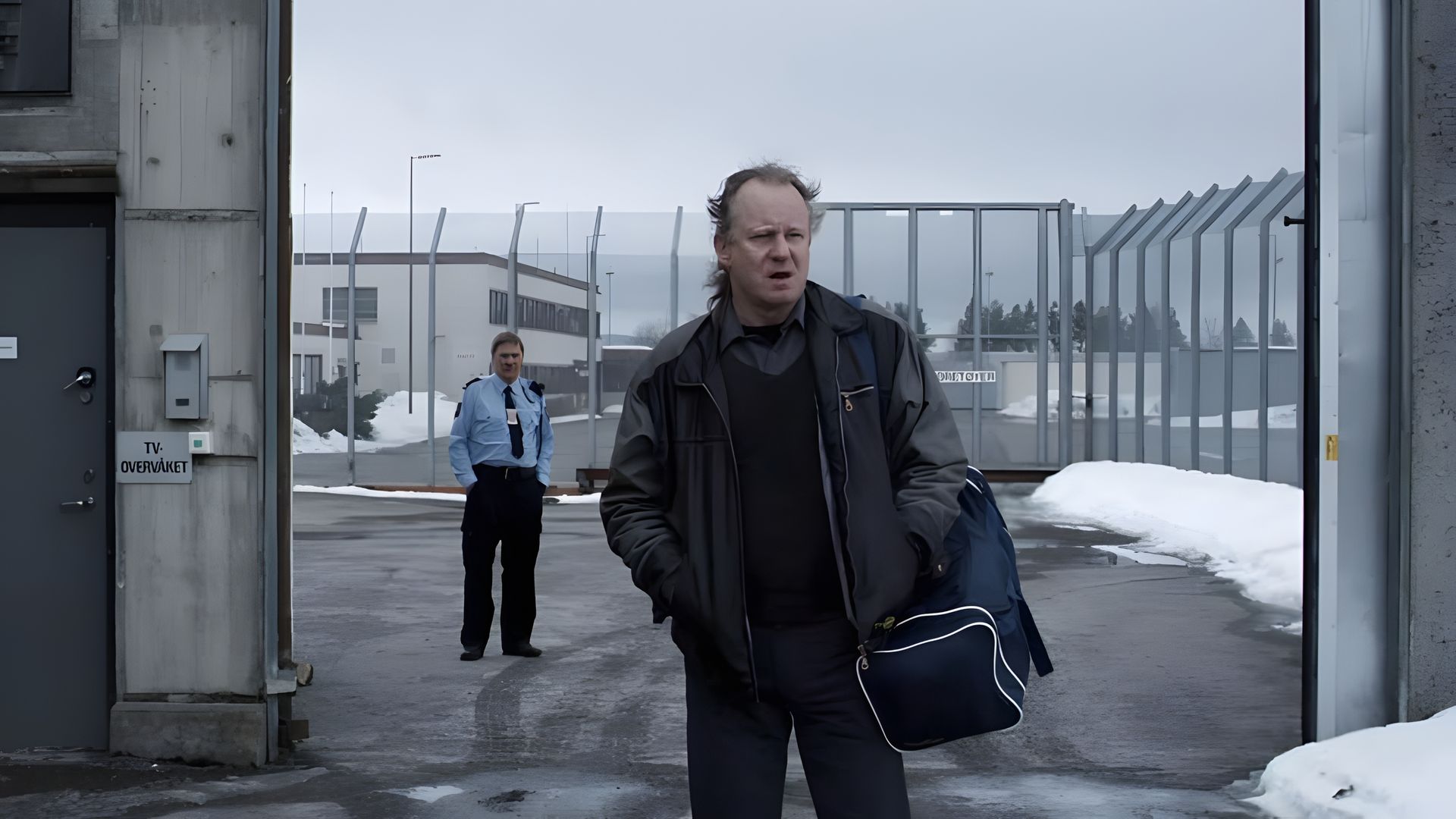 Stellan Skarsgård che porta una borsa esce dai cancelli della prigione, con una guardia sullo sfondo, edifici e neve visibili oltre la recinzione