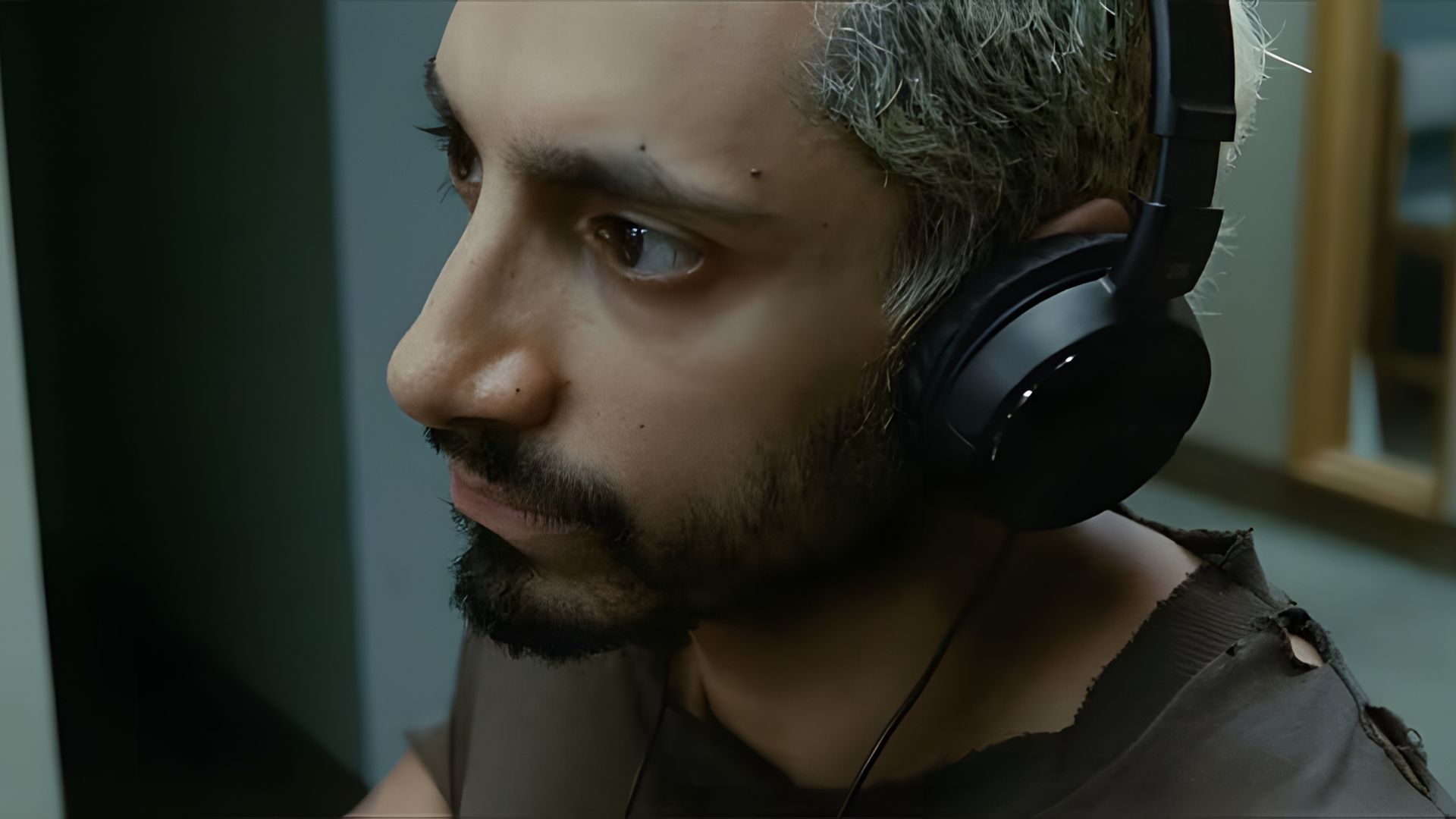 Riz Ahmed indossa le cuffie con un'espressione seria, seduto in interni, vestito con una maglietta strappata