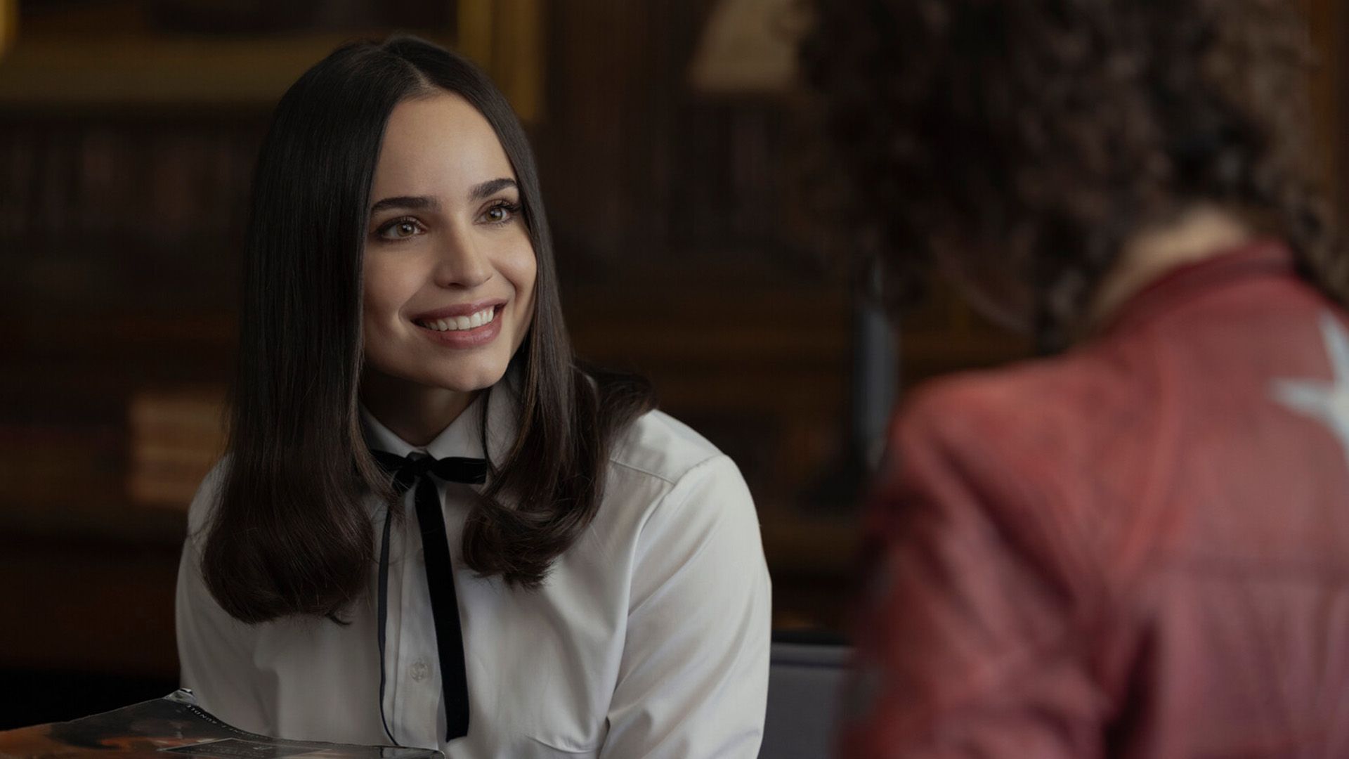 Sofia Carson in una camicetta bianca con un fiocco nero al collo sorride ampiamente