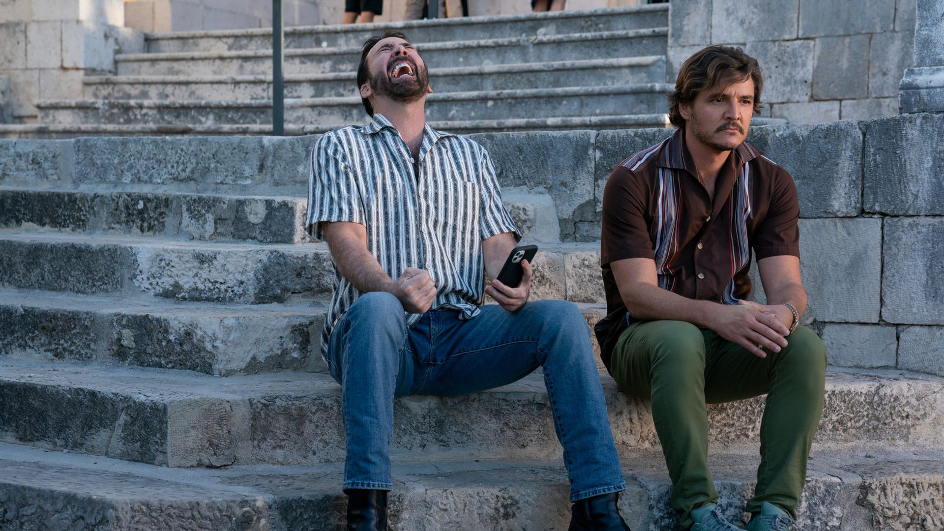 Nicolas Cage e Pedro Pascal seduti sui gradini di pietra: uno ride con un telefono in mano, l'altro guarda pensieroso di lato