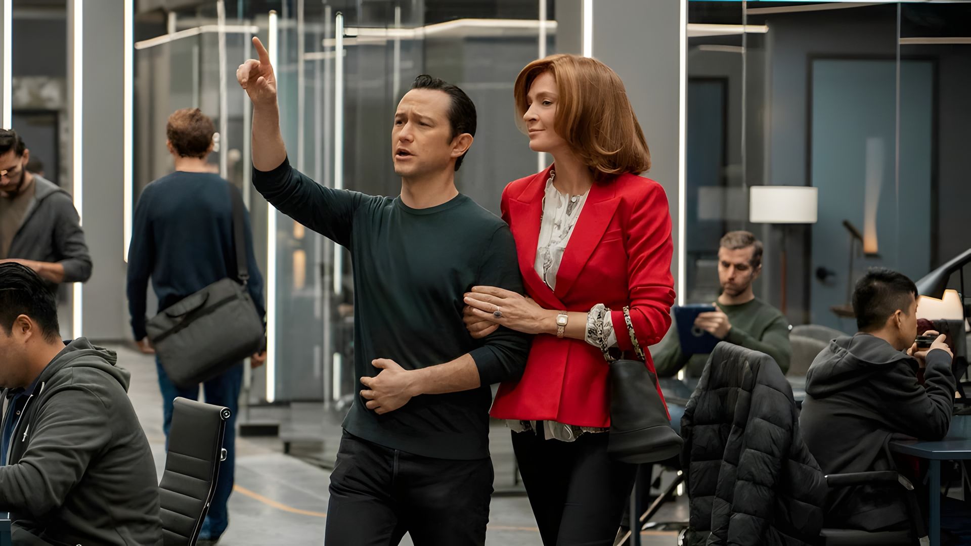 Joseph Gordon-Levitt in abiti scuri punta in avanti mentre Uma Thurman in giacca rossa si aggrappa al suo braccio