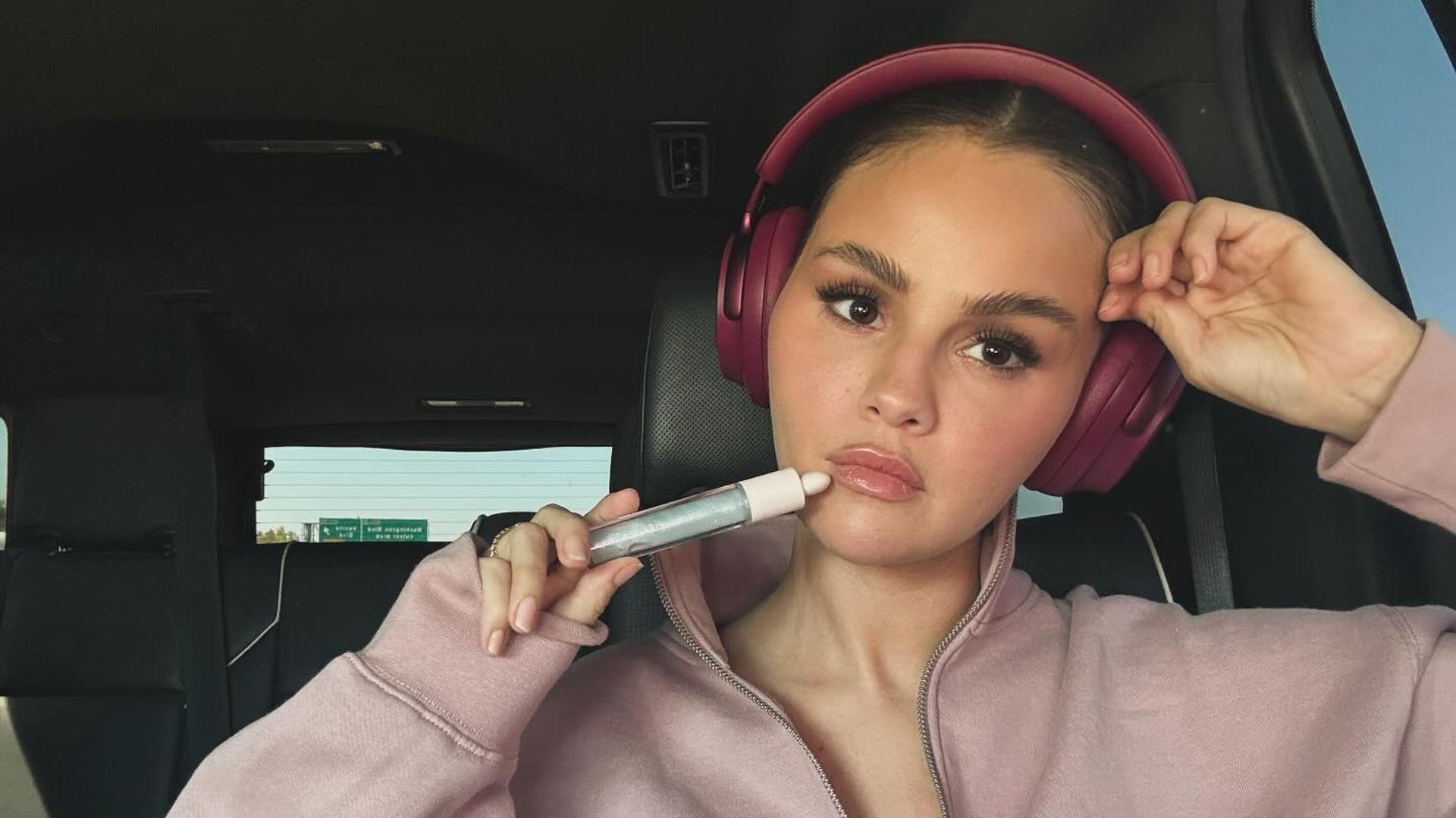 Selena Gomez in una felpa rosa e cuffie seduta in macchina, tiene un prodotto cosmetico vicino al viso