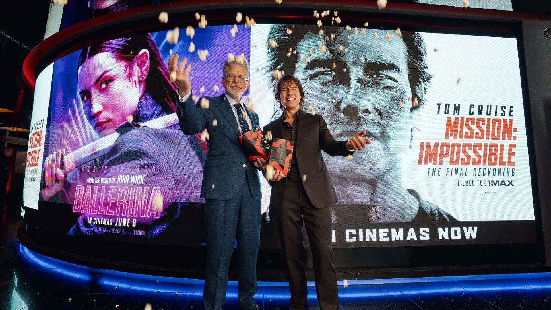 Due uomini in giacca e cravatta posano con scatole di popcorn sullo sfondo di poster cinematografici per Ballerina e Mission: Impossible
