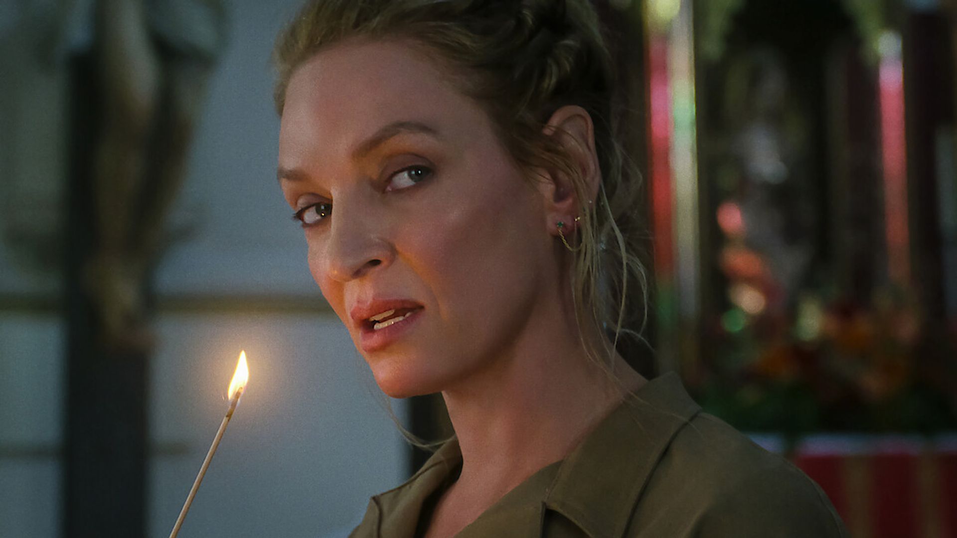 Uma Thurman con i capelli raccolti tiene una candela accesa e guarda di lato con uno sfondo leggermente sfocato