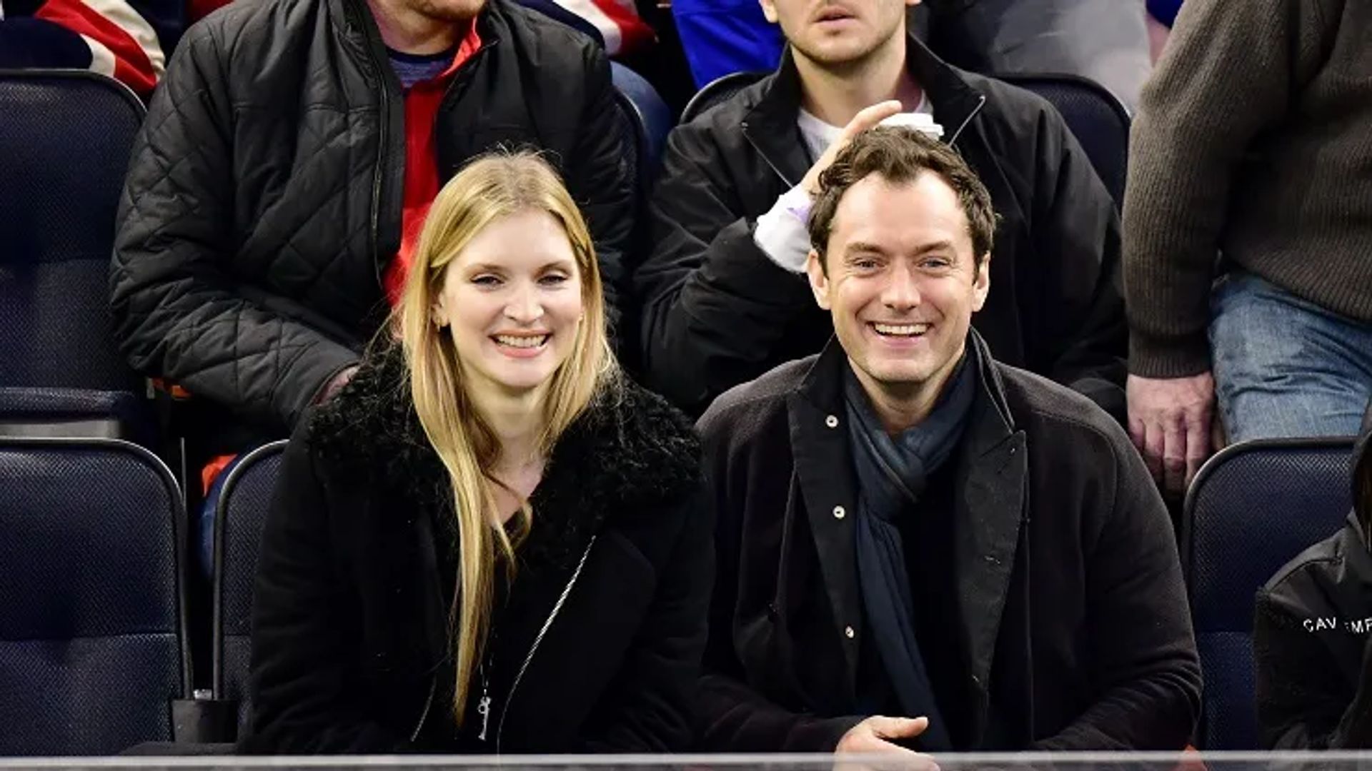 Jude Law e Phillipa Coan seduti fianco a fianco sugli spalti, sorridenti e che guardano verso la telecamera