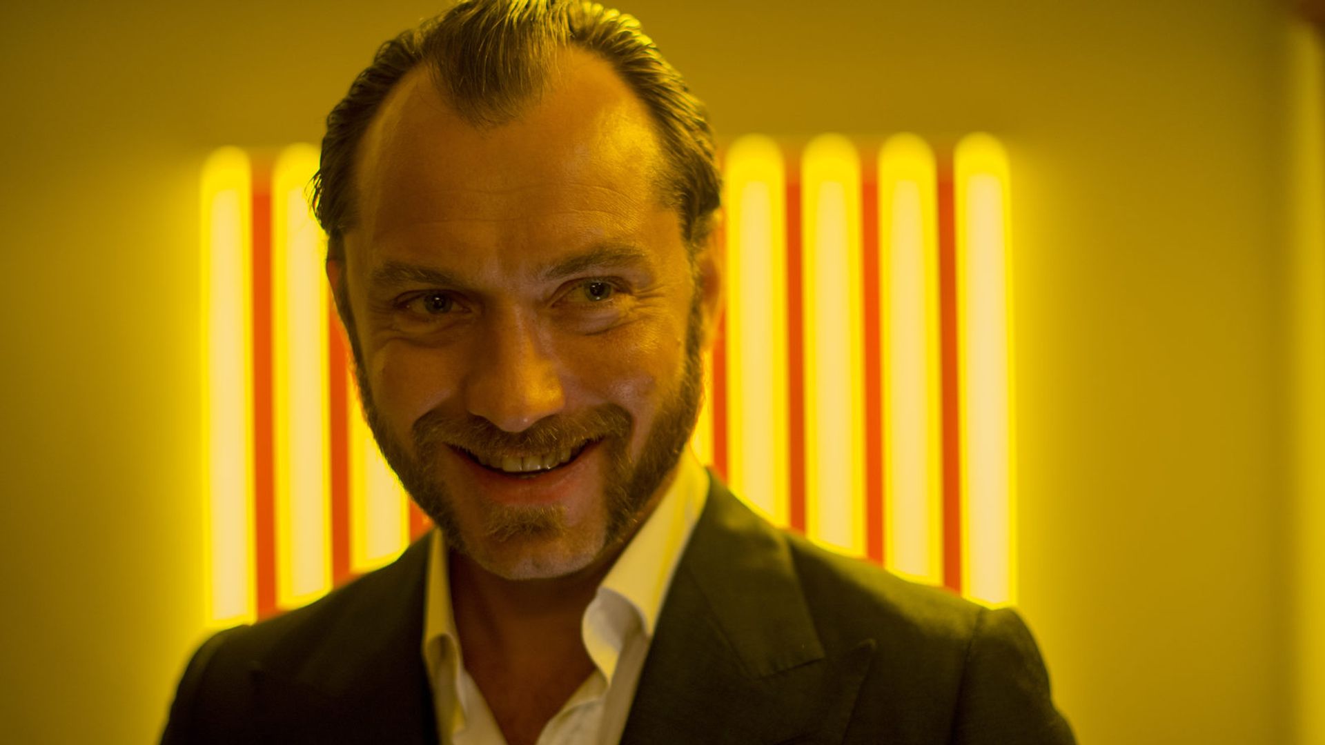 Jude Law in giacca sorride su uno sfondo di strisce di luce gialla brillante