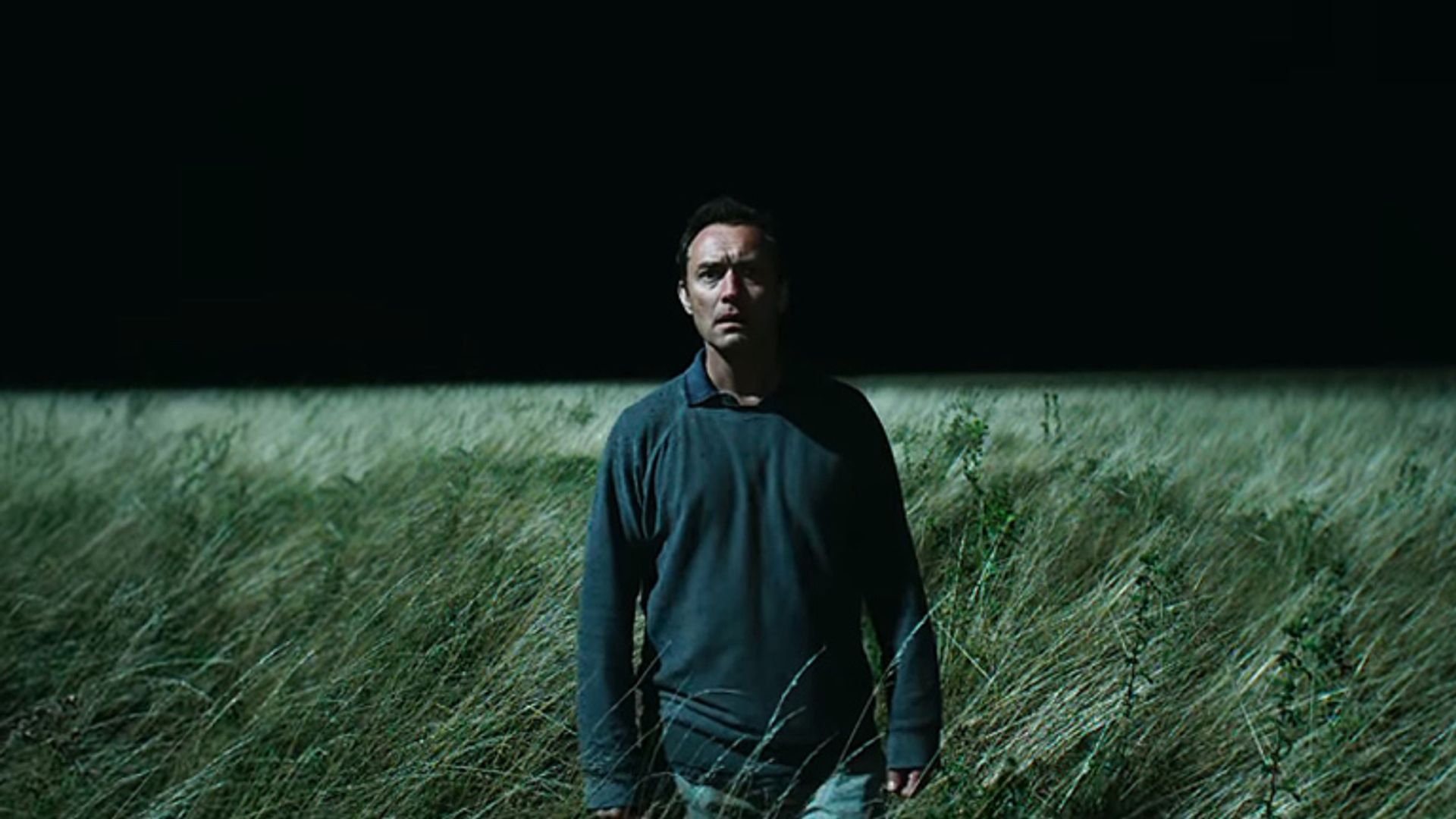 Jude Law in abiti scuri in piedi in un campo di erba alta di notte, illuminato da una luce intensa