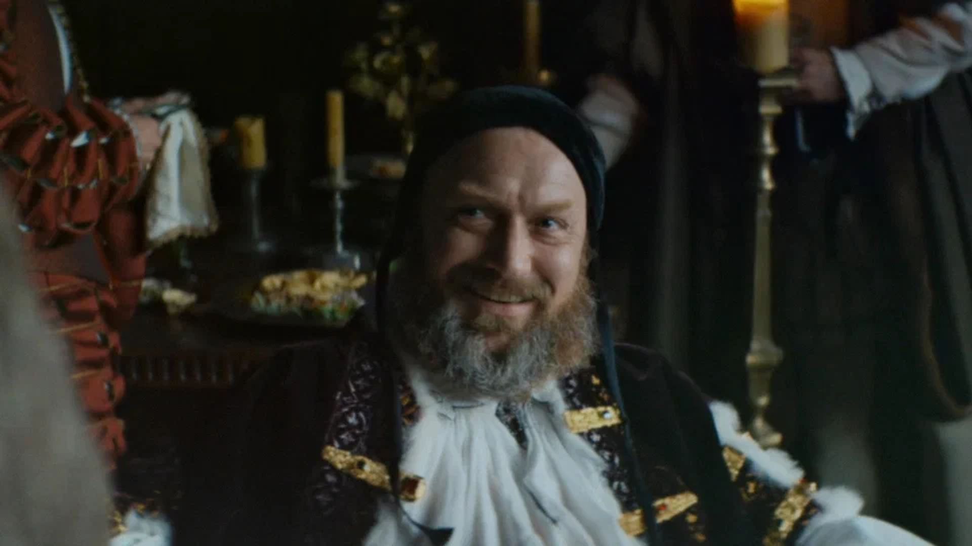 Jude Law in costume medievale con la barba sorride mentre siede a un tavolo festivo illuminato dalle candele